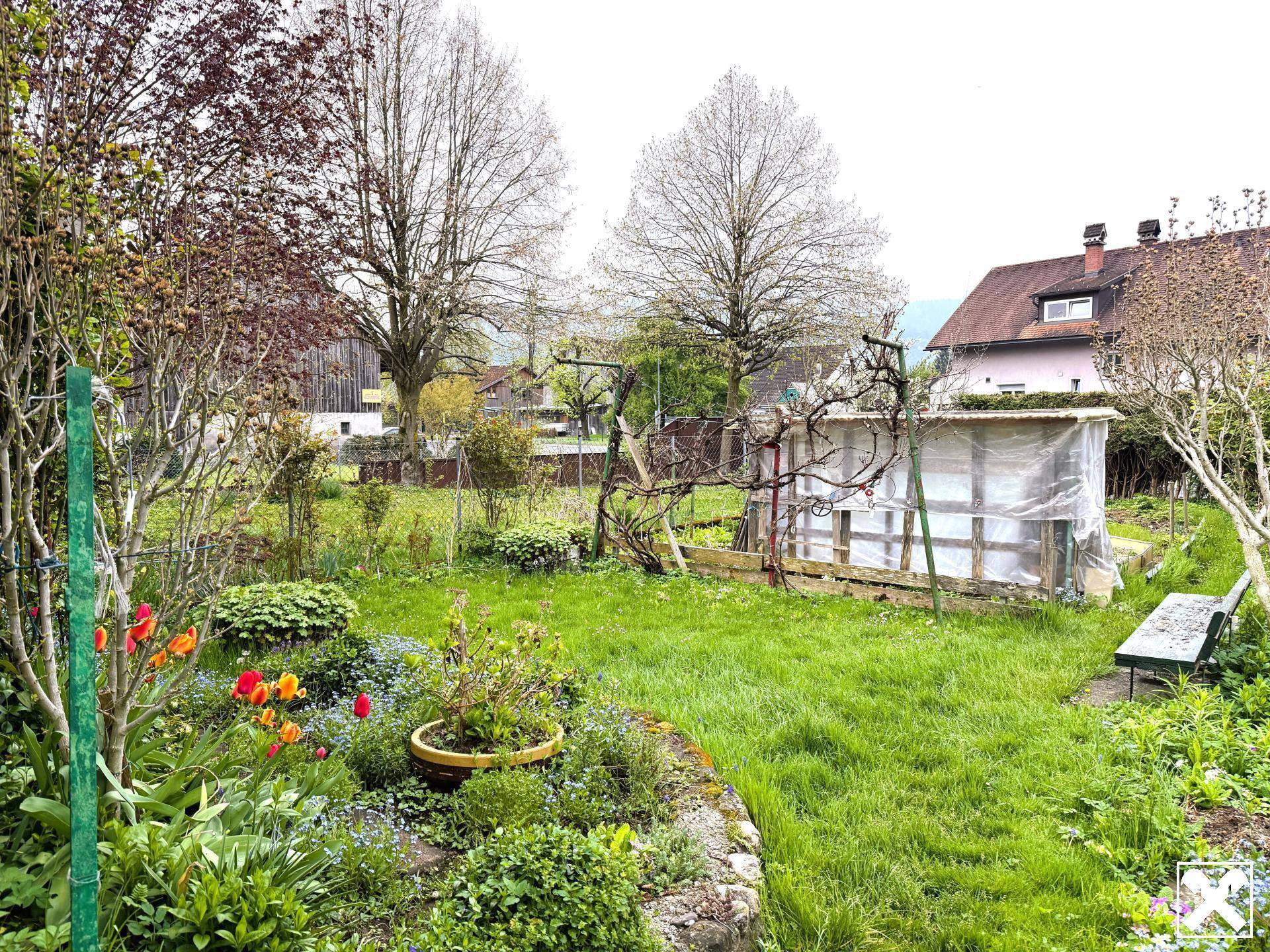 Großer Garten mit Rasenfläche, Blumenbeeten und einem kleinen Gewächshaus.