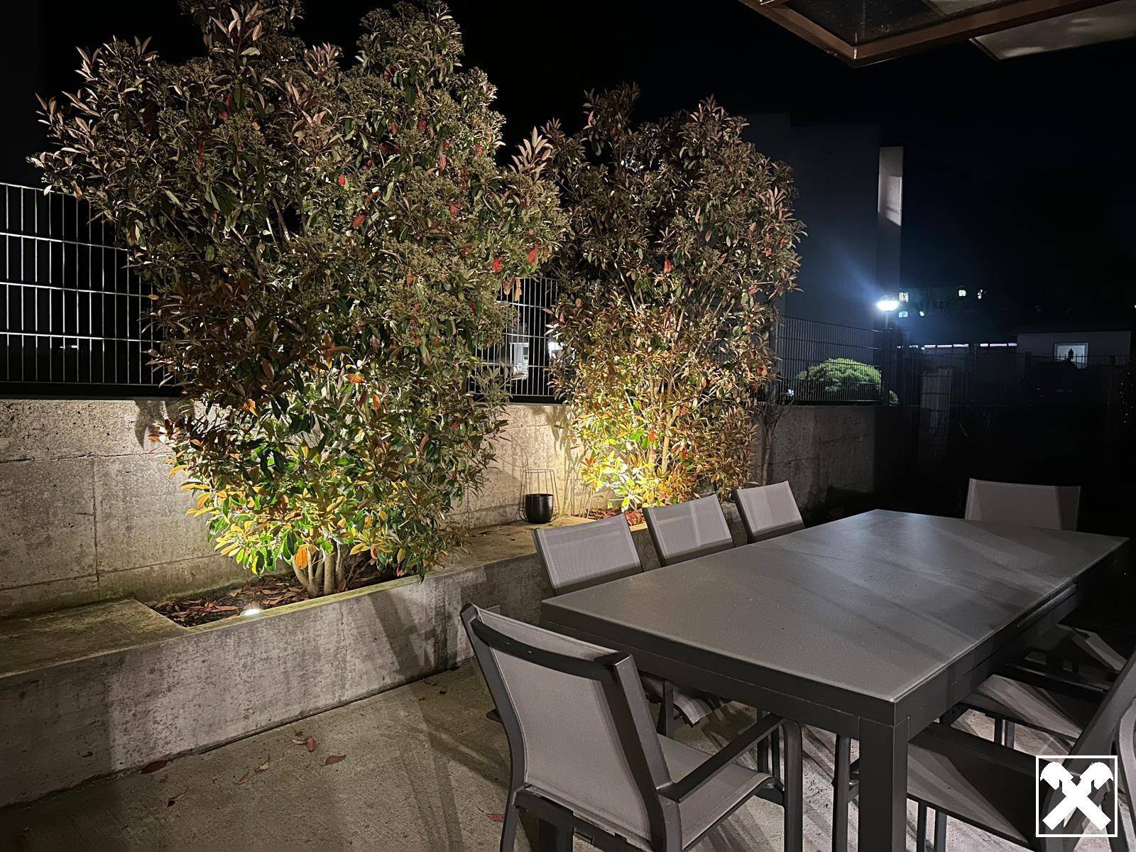Beleuchtete Terrasse mit Esstisch und Stühlen, umgeben von bepflanzten Beeten.