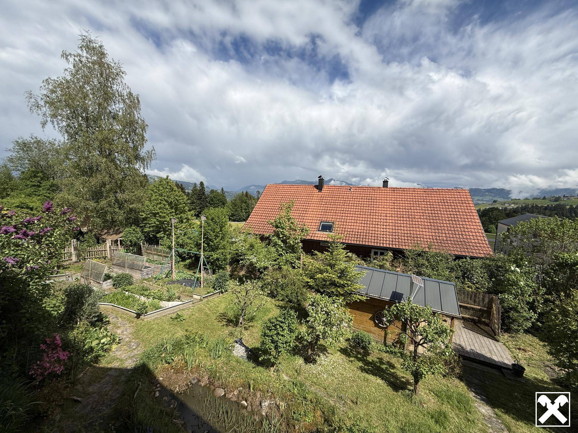 Weitläufiger Blick auf das Haus mit Garten, Hochbeeten und der umliegenden grünen Berglandschaft.