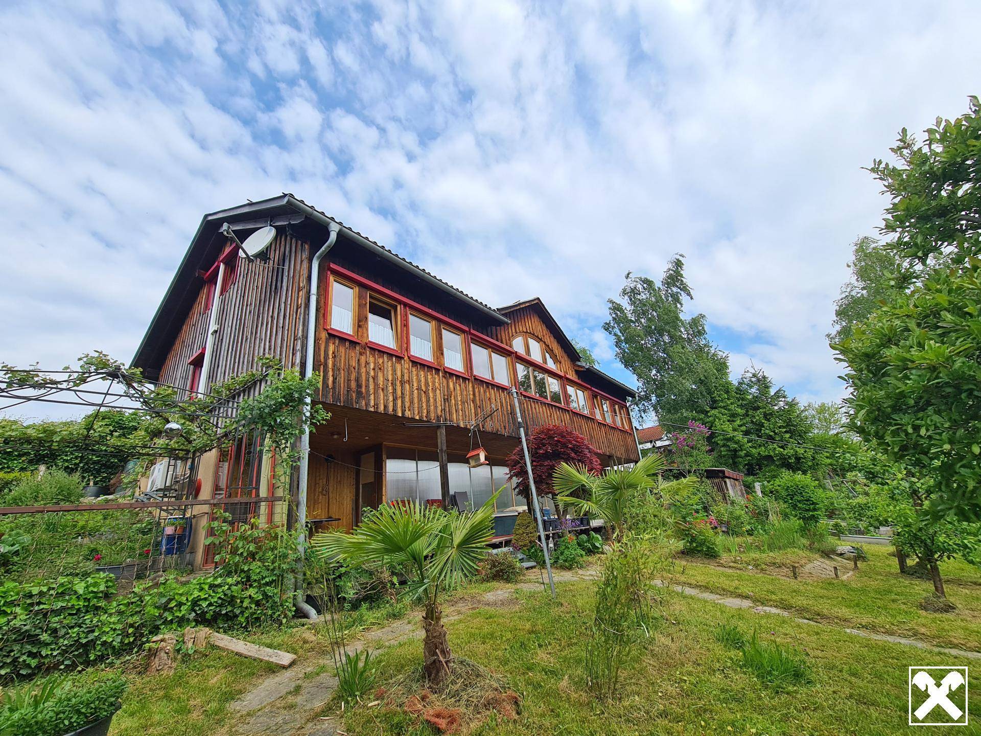 Rustikales Holzhaus mit großen Fenstern, umgeben von üppiger Vegetation unter blauem Himmel.
