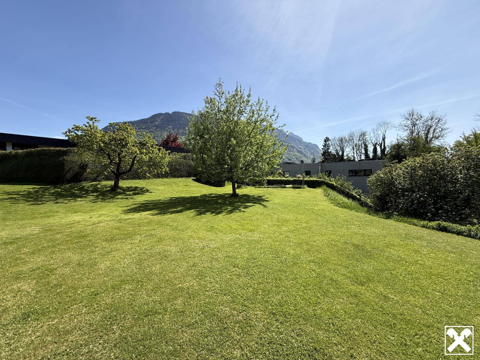 Weitläufiger, gepflegter Garten mit Rasenflächen und Bäumen, umgeben von grüner Natur und Bergen.