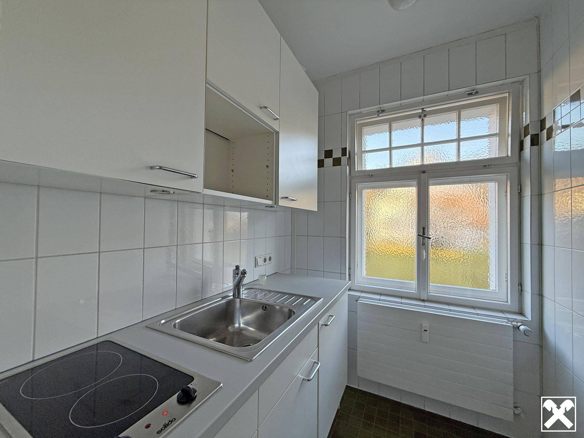 Kleine Küche mit weißen Schränken, Spüle und Kochfeld, komplett gefliesten Wänden und einem Fenster.