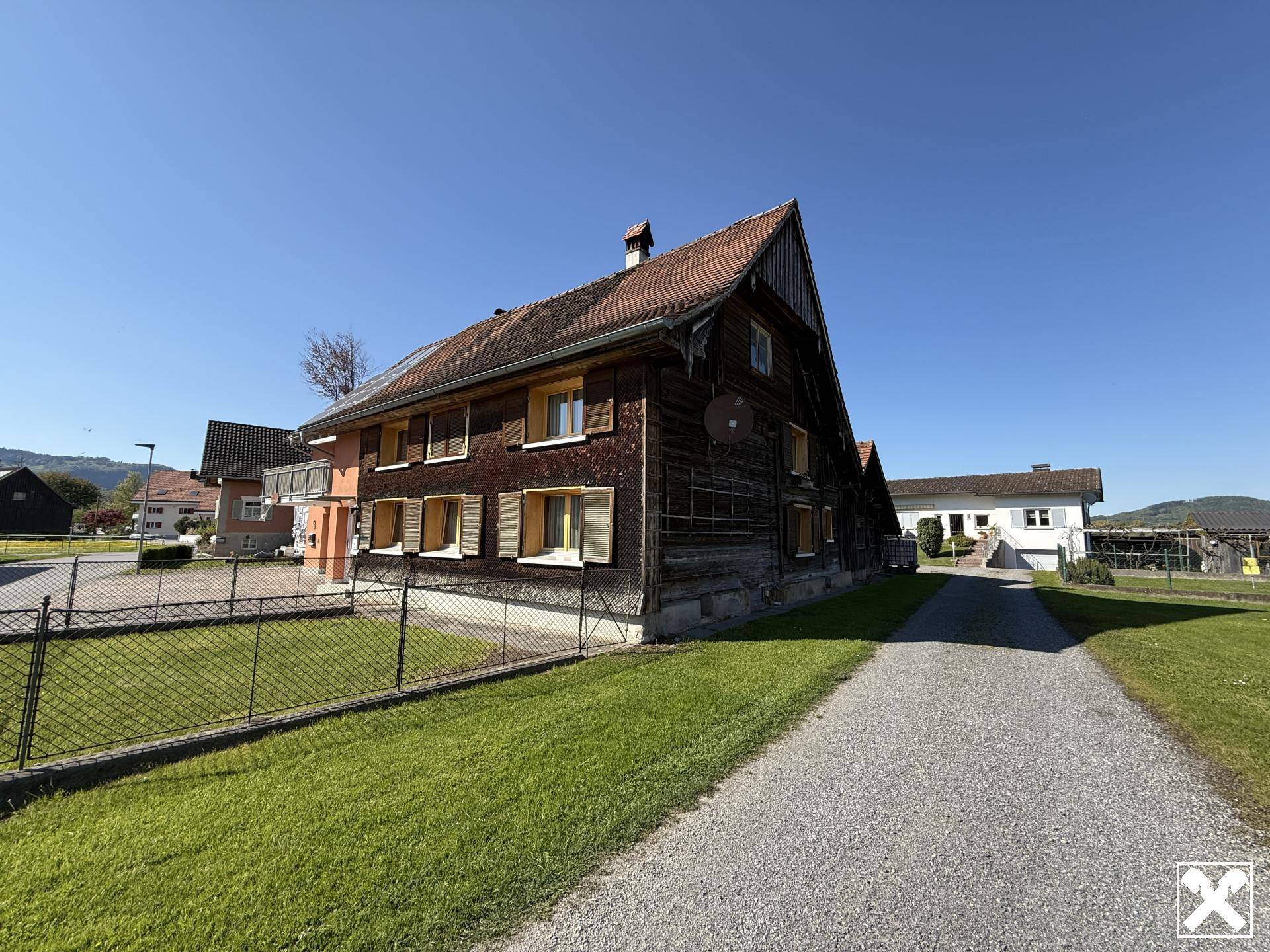 Seitenansicht des Hauses mit Holzwänden, Kiesweg und weitläufiger Rasenfläche unter blauem Himmel.
