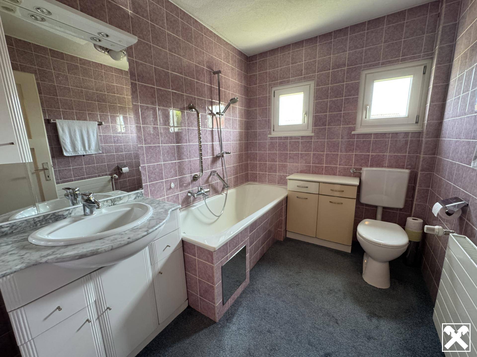 Badezimmer mit lila Fliesen, Badewanne, Waschbecken und zwei Fenstern.