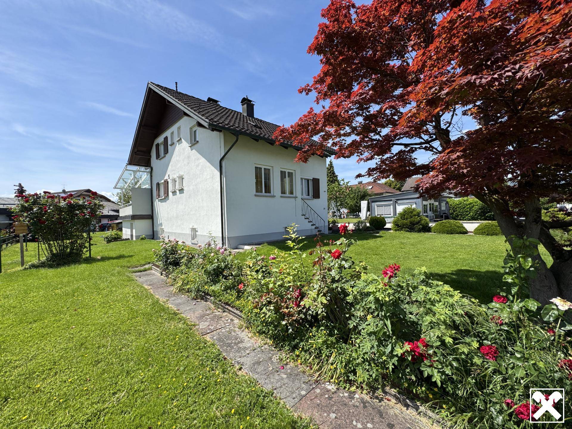 Seitenansicht des Hauses mit blühenden Rosenbeeten und einem großen roten Ahornbaum im Garten.