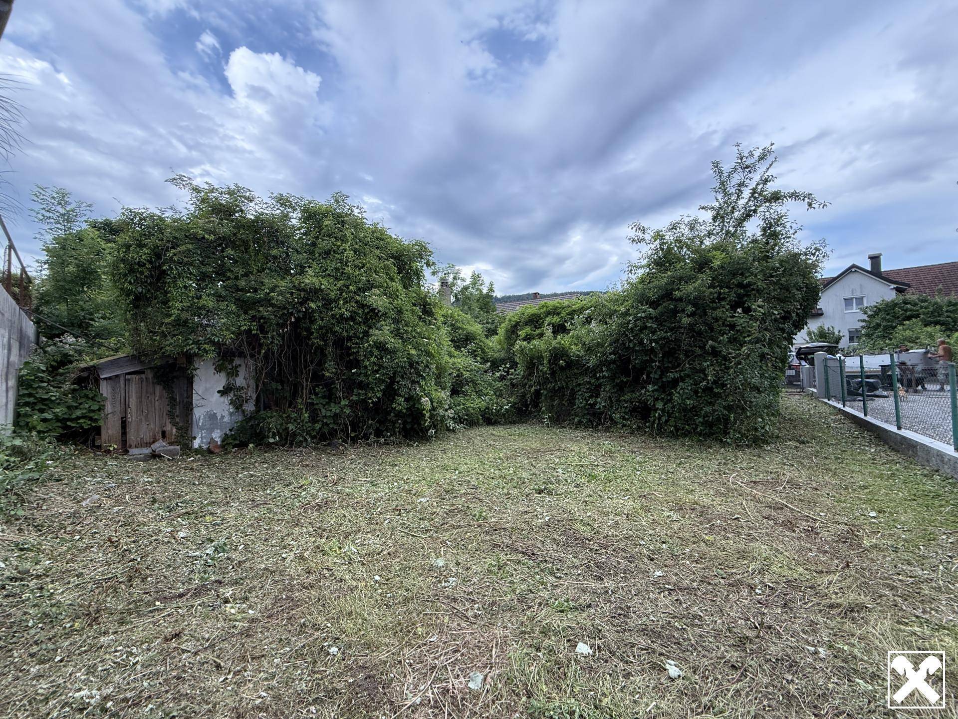 Verwildertes Grundstück mit einem kleinen, verfallenen Schuppen und dichter Bepflanzung.