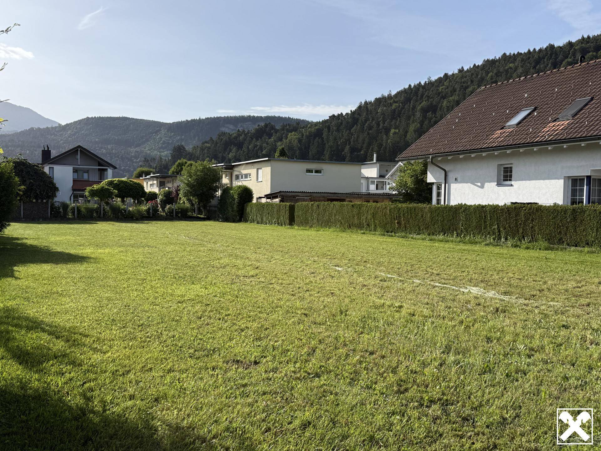 Großzügige Rasenfläche eines Grundstücks, umgeben von Häusern und grünen Hügeln unter blauem Himmel.