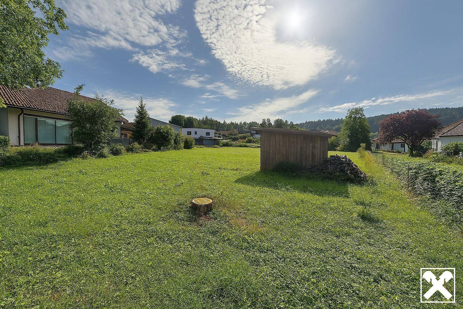 Sonnige Grünfläche mit einem Holzschuppen und Baumstumpf, umgeben von ländlicher Bebauung.