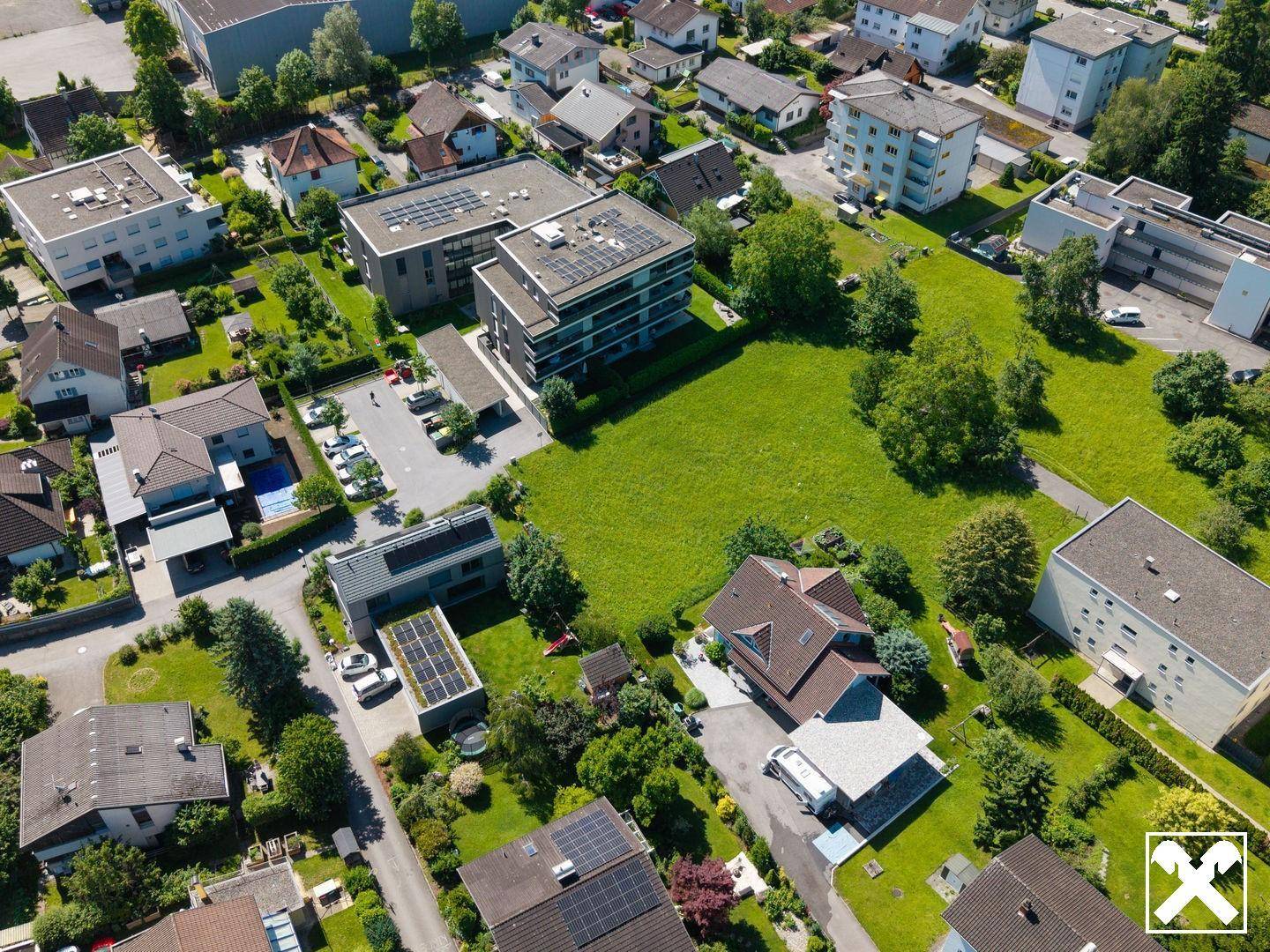 Luftaufnahme eines Wohngebiets mit einem großen grünen Grundstück, umgeben von Häusern und modernen Gebäuden mit Solaranlagen.