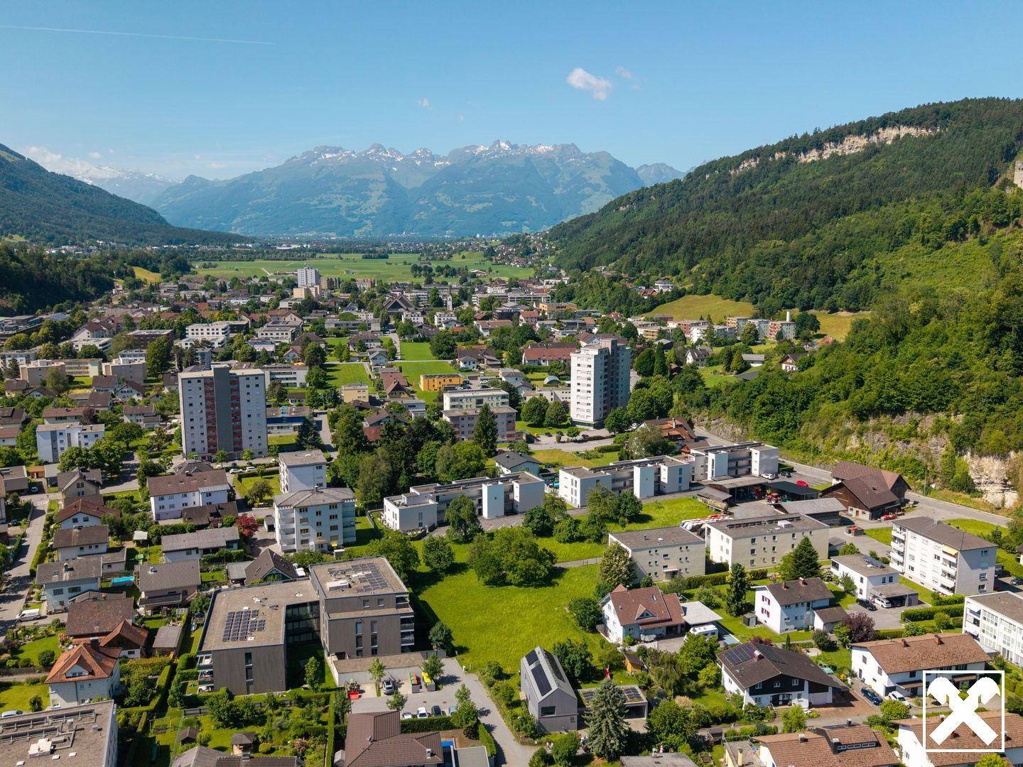 Weitläufige Luftaufnahme einer Wohnsiedlung mit grünen Hügeln und schneebedeckten Bergen im Hintergrund unter blauem Himmel.