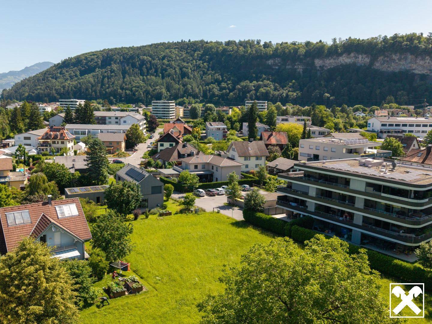 Luftaufnahme eines Wohngebiets mit einem großen grünen Grundstück im Vordergrund und Bergen im Hintergrund.