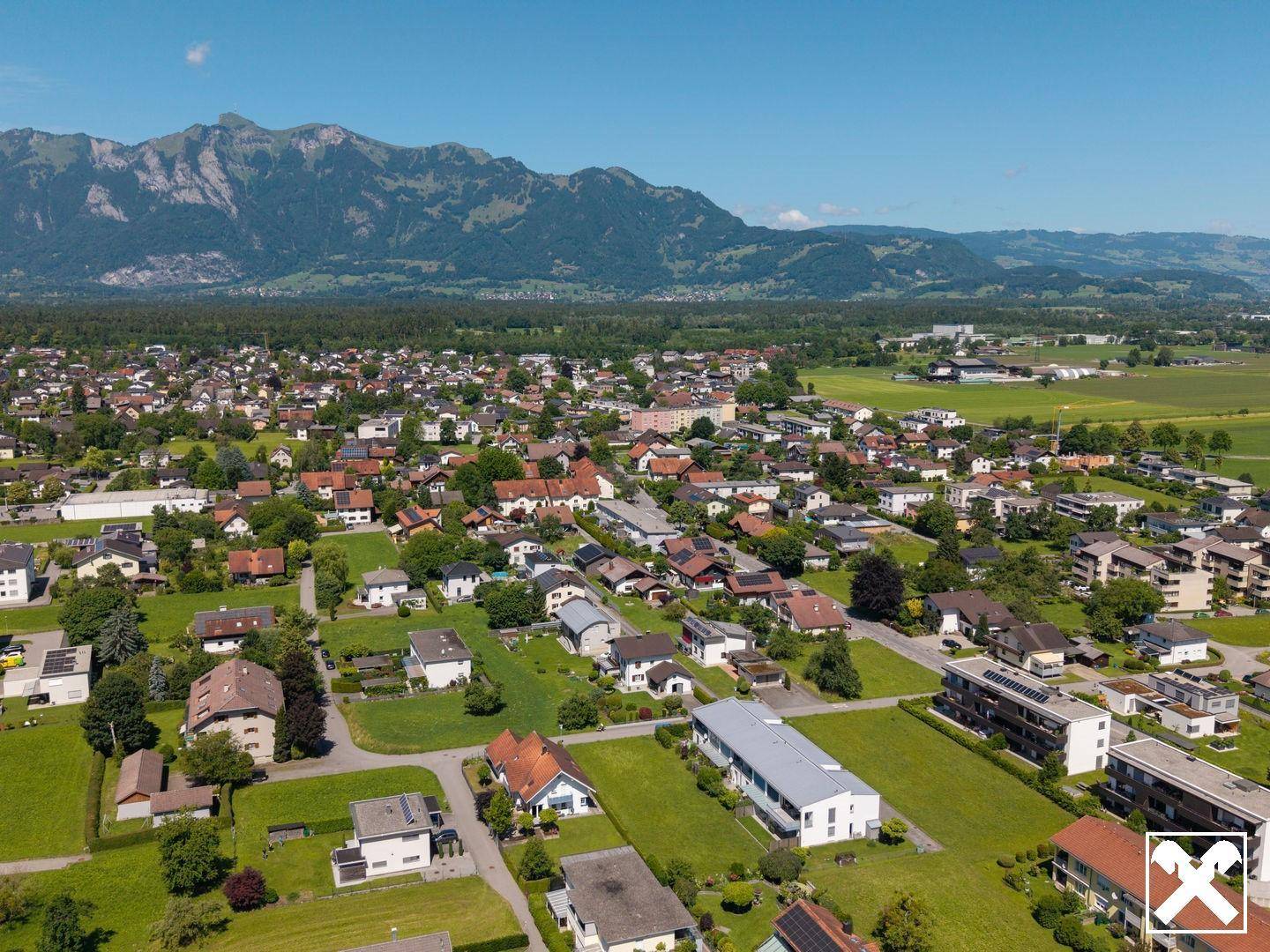 Farbige Luftaufnahme einer weitläufigen Wohnsiedlung mit grünen Feldern und imposantem Bergpanorama.