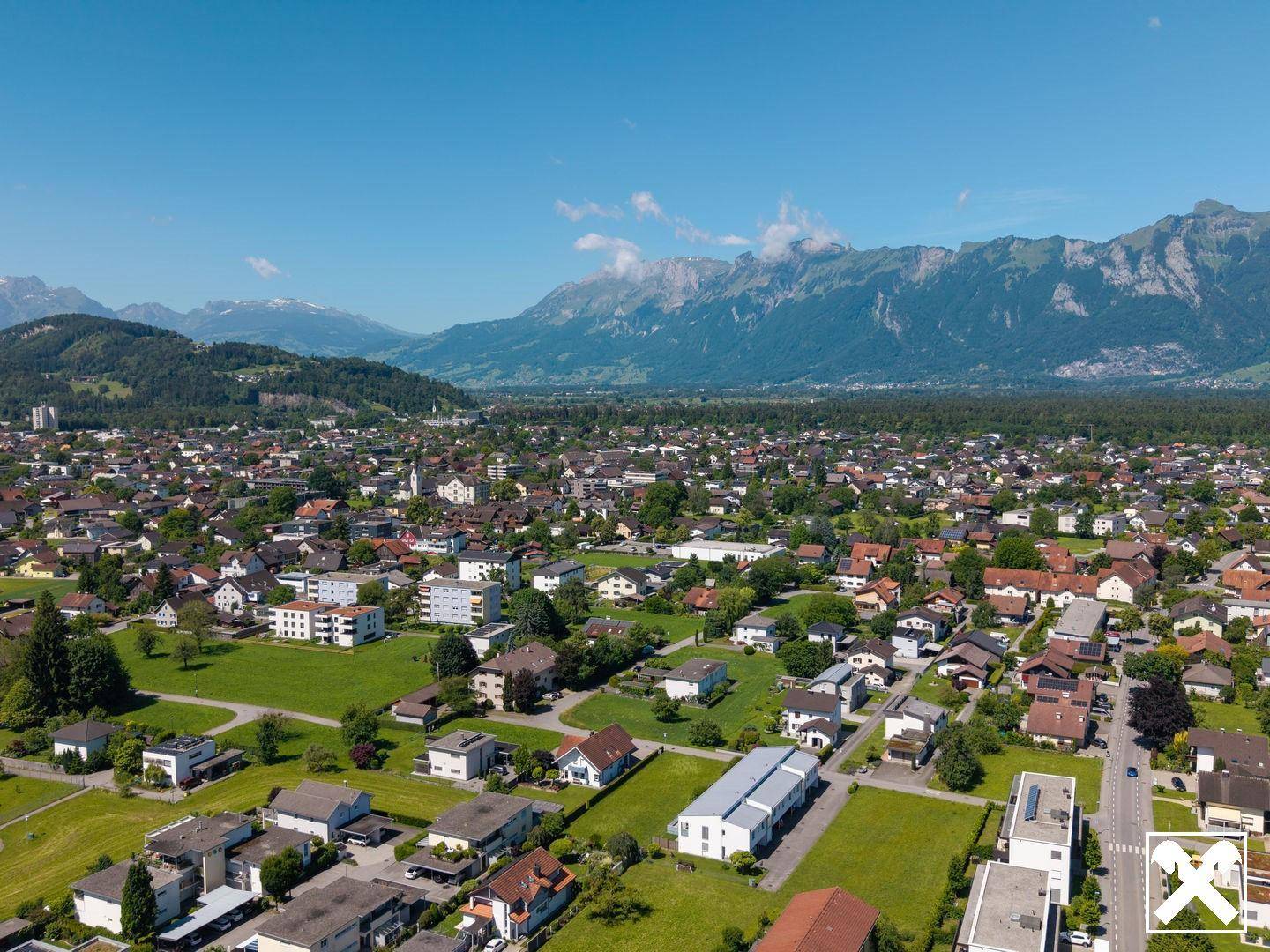 Panoramische Luftaufnahme einer Wohnsiedlung mit weitläufigen Grünflächen und einer majestätischen Bergkette.