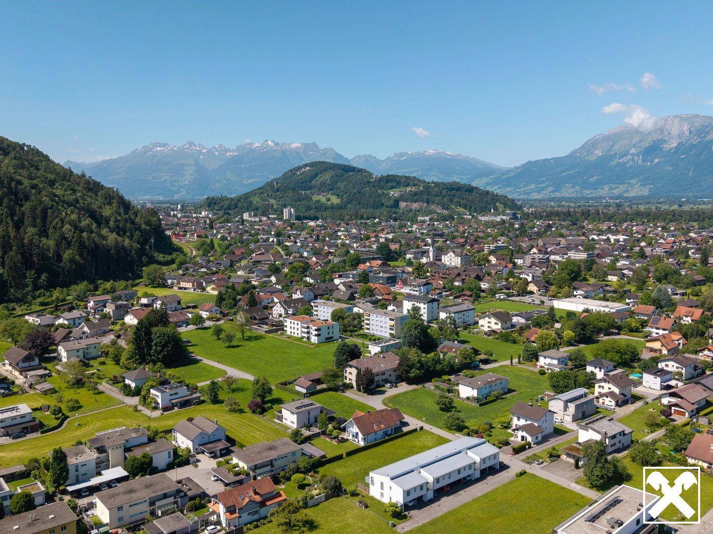 Weitwinkel-Luftaufnahme einer ausgedehnten Wohngegend, eingebettet zwischen grünen Hügeln und hohen Bergen.