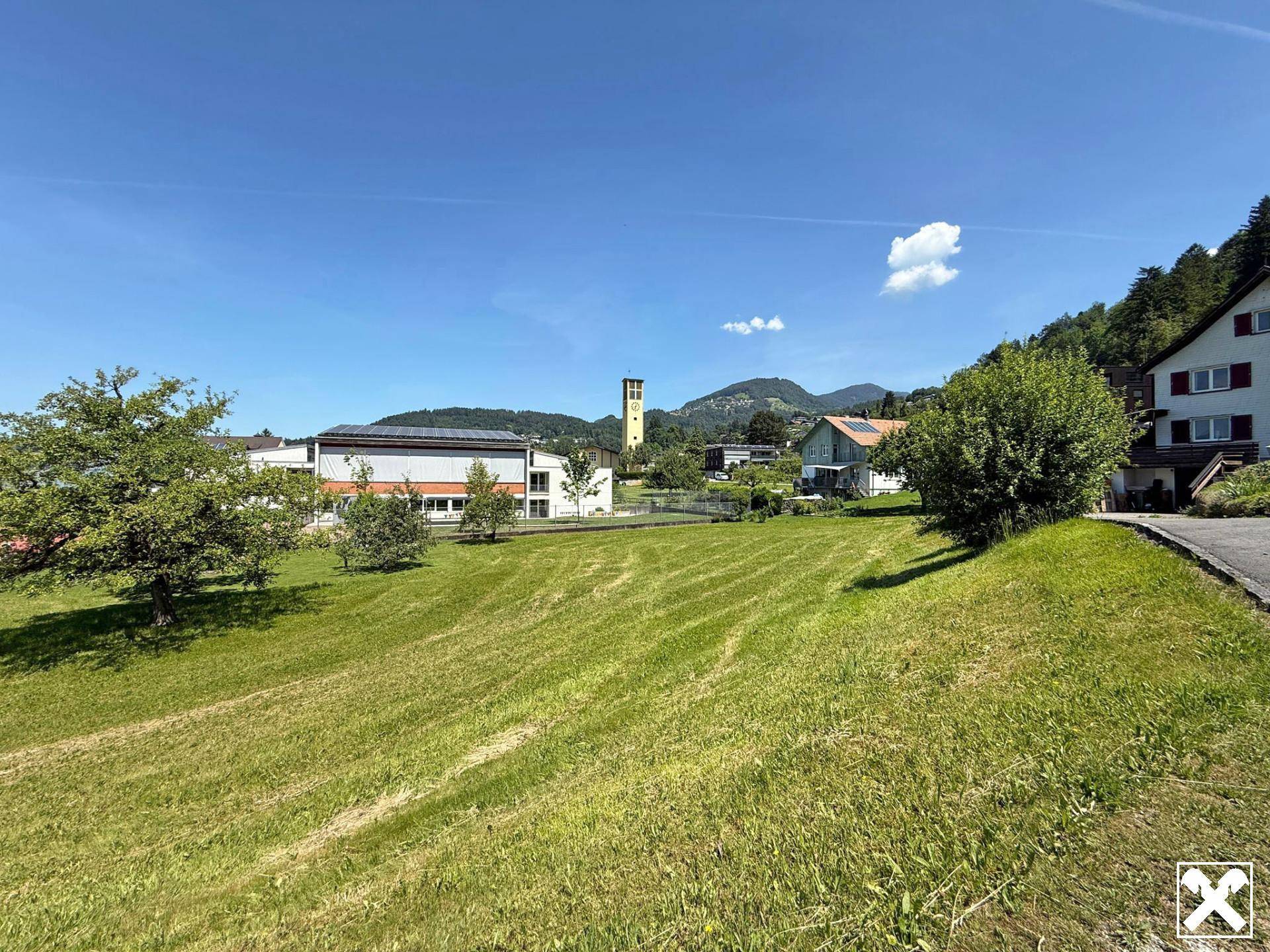 Weitläufige Wiese mit Blick auf eine Kirche, umliegende Häuser und ferne Berge.