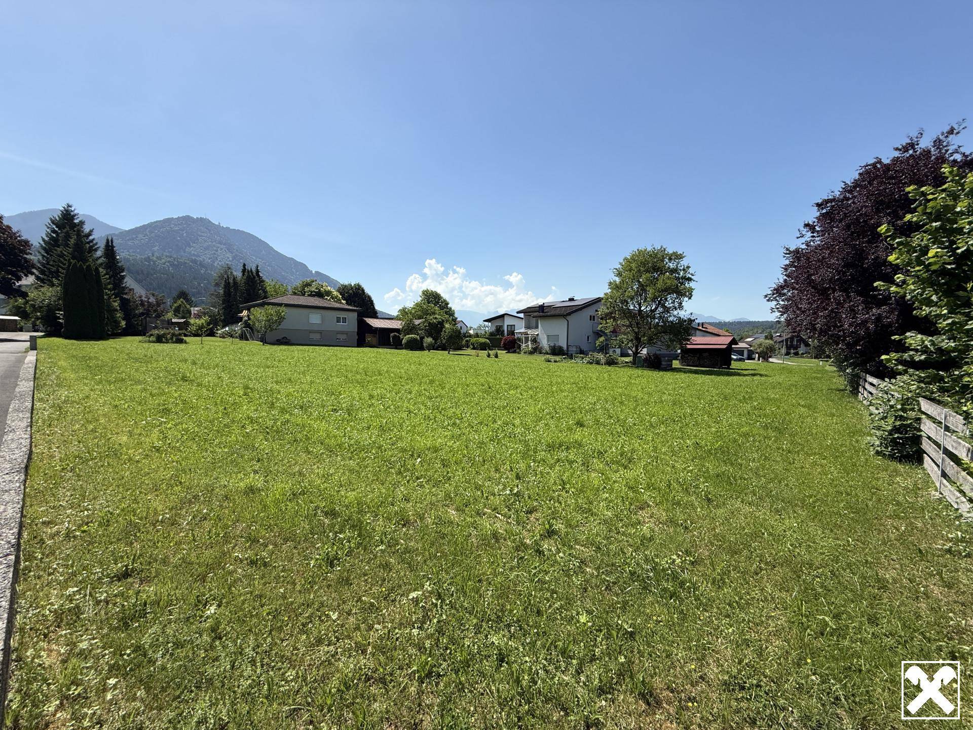 Sonniger Blick über ein großes grünes Grundstück mit angrenzenden Häusern und Bergpanorama im Hintergrund.