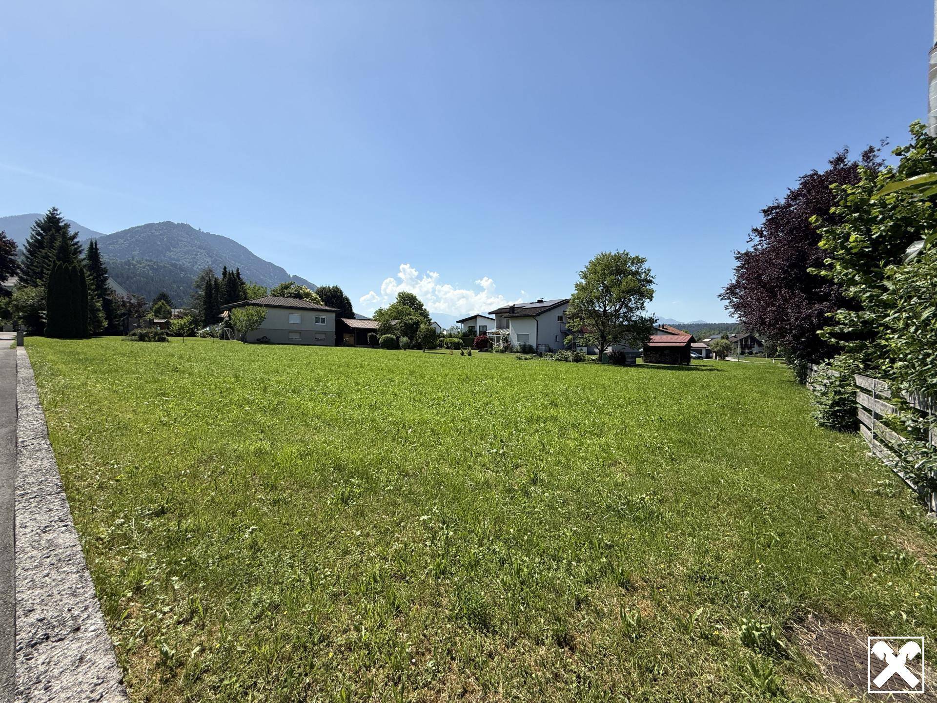 Ausgedehntes grünes Grundstück mit viel Sonnenschein, umgeben von Wohnbebauung und alpiner Landschaft.