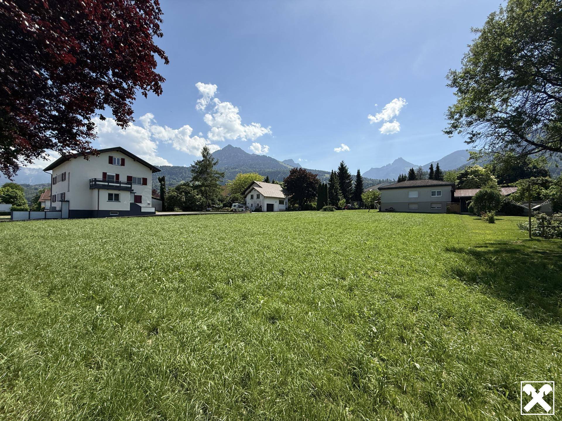 Weitläufige Grünfläche mit beeindruckendem Bergblick und umliegenden Wohnhäusern unter strahlend blauem Himmel.