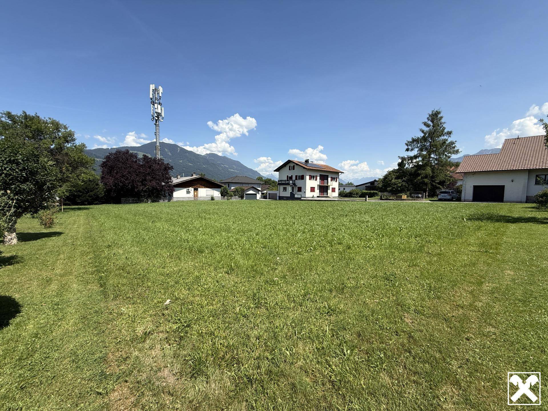 Großes, sonniges Grundstück mit grüner Wiese, umgeben von Häusern und einem weiten Blick auf die Berge.