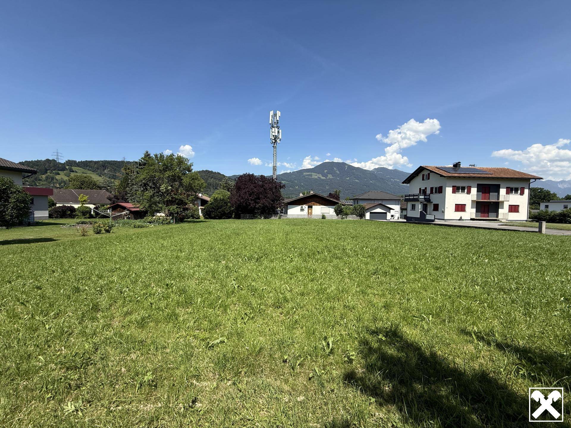 Ausgedehntes grünes Grundstück mit viel Sonnenschein, umgeben von Wohnbebauung und alpiner Landschaft.