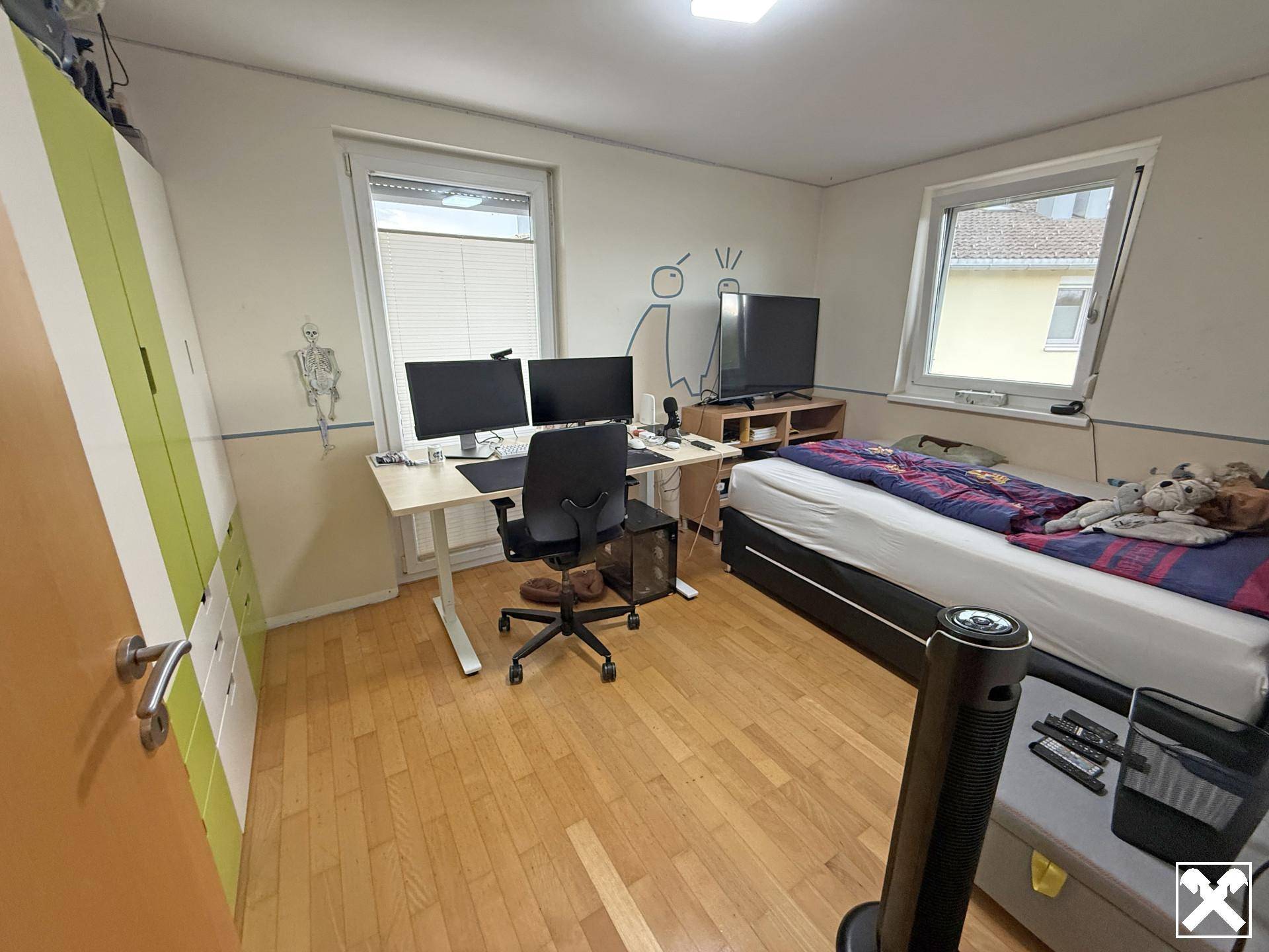 Kinder- oder Arbeitszimmer mit Holzboden, Schreibtisch, Bett und zwei Fenstern.