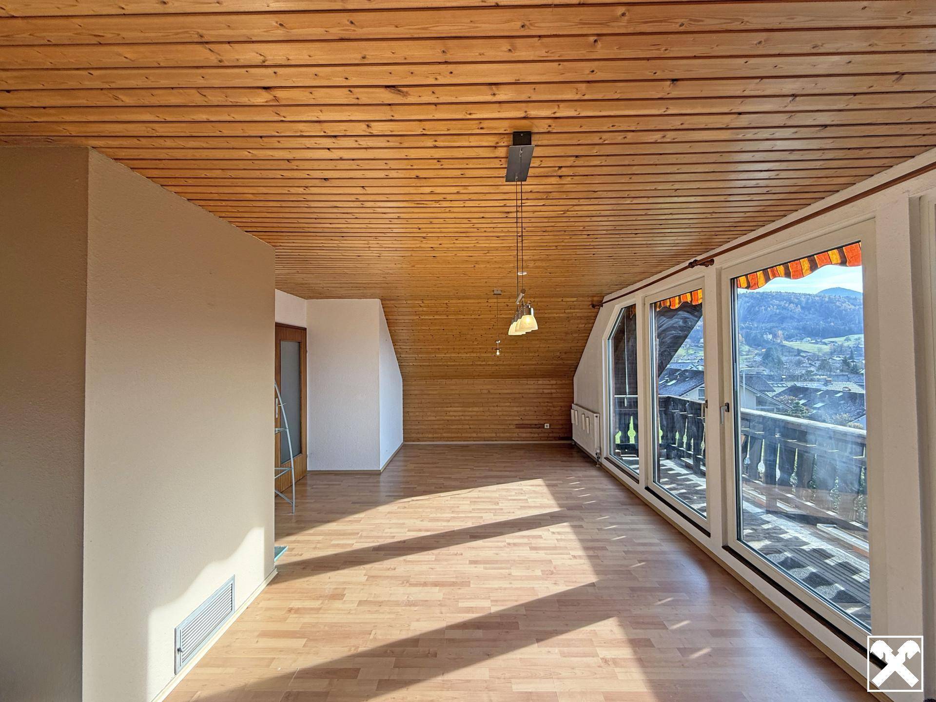 Geräumiger Wohnbereich im Dachgeschoss mit Holzdecke, großen Fenstern und Zugang zum Balkon.