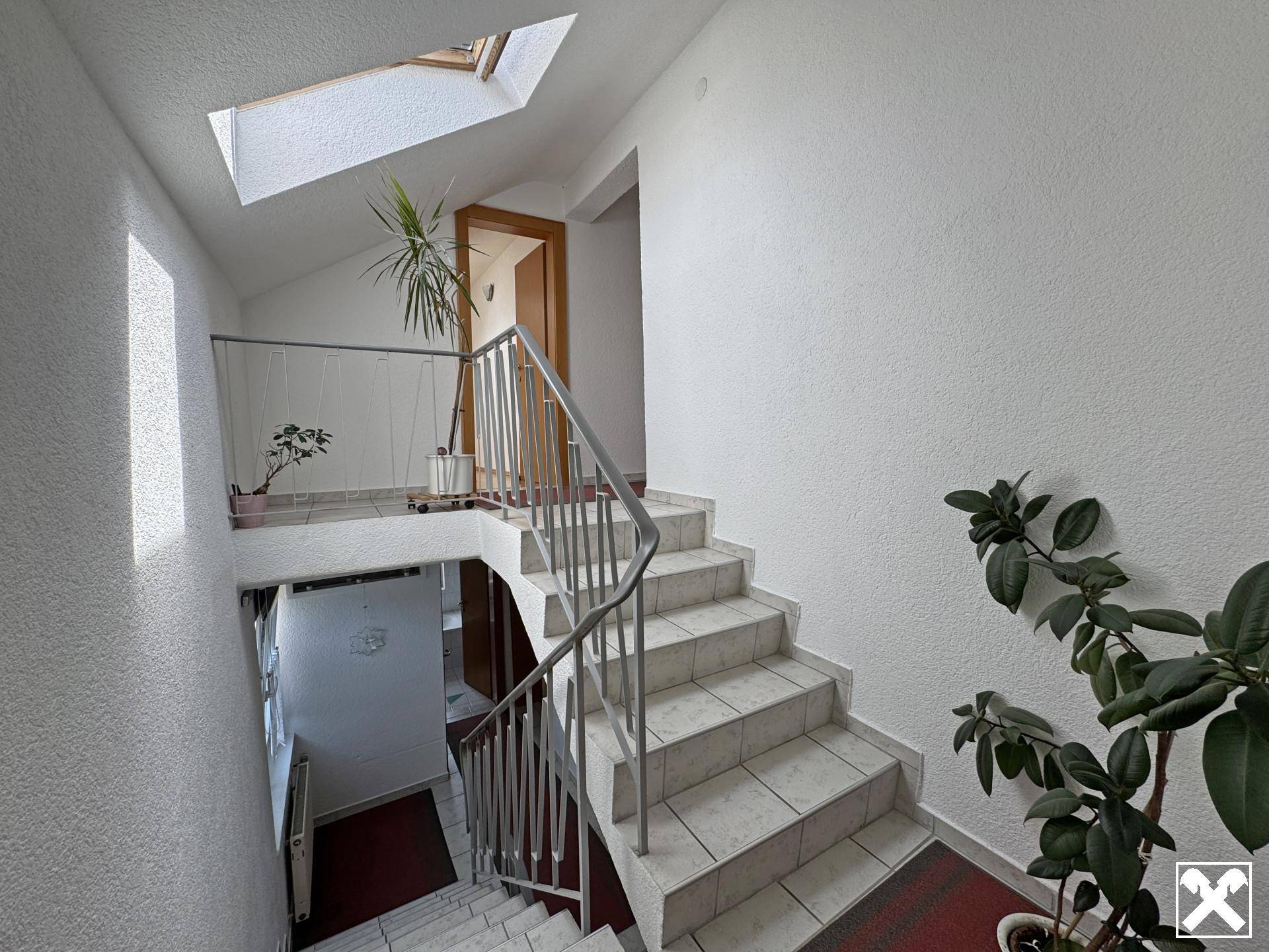 Heller Treppenaufgang mit Fliesenboden, Metallgeländer und natürlichem Licht durch ein Dachfenster.