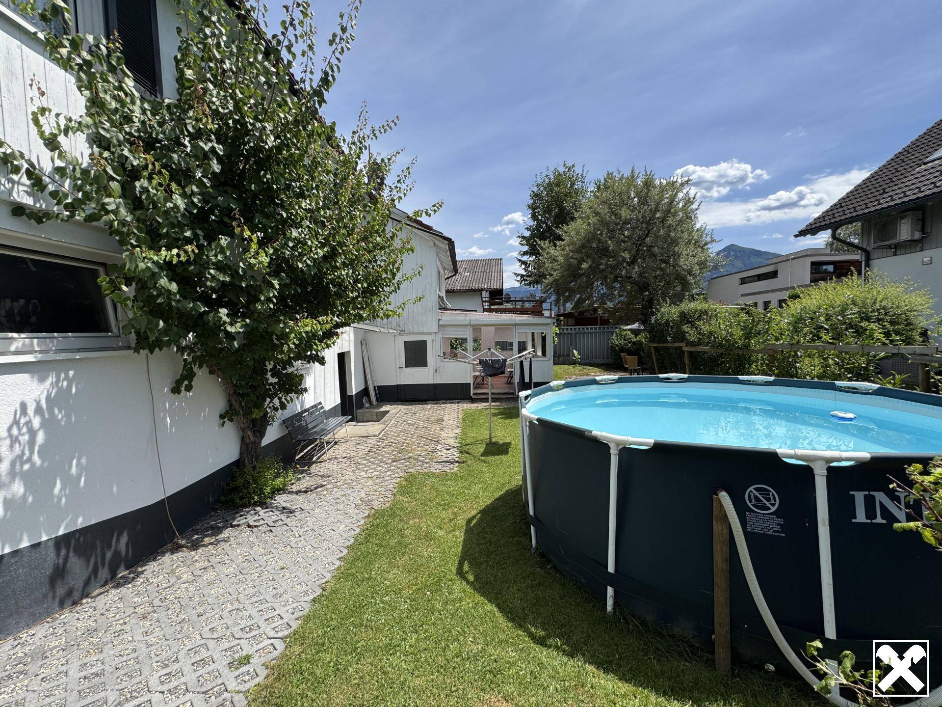Seitlicher Gartenbereich mit Aufstellpool, gepflastertem Weg und Blick auf die Berge.