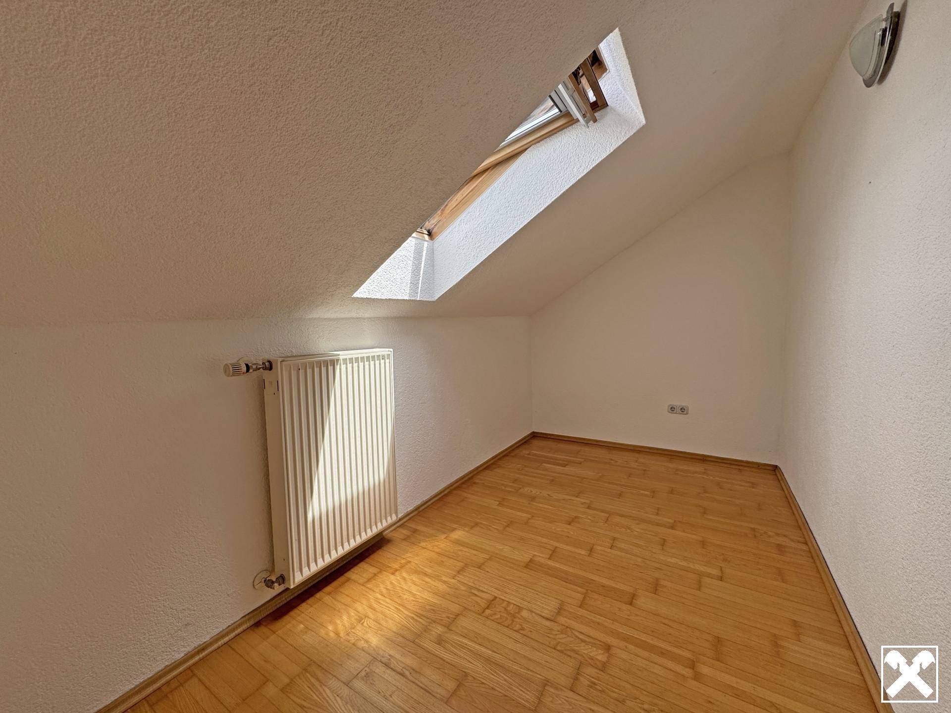 Helles Zimmer im Dachgeschoss mit Holzparkettboden, Dachschräge und einem Dachfenster.