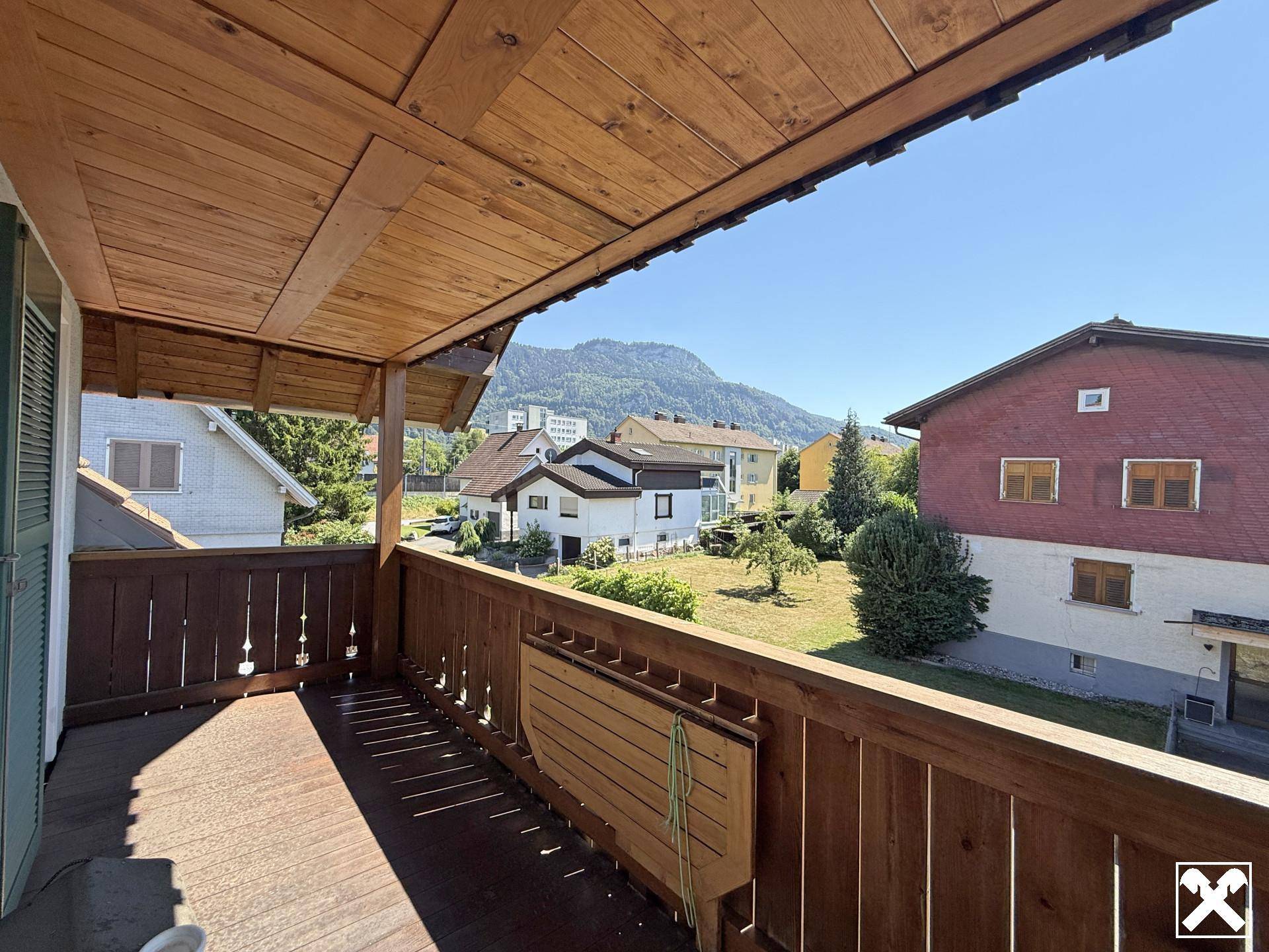 Holzbalkon mit Bergblick und umliegender grüner Landschaft unter strahlend blauem Himmel.