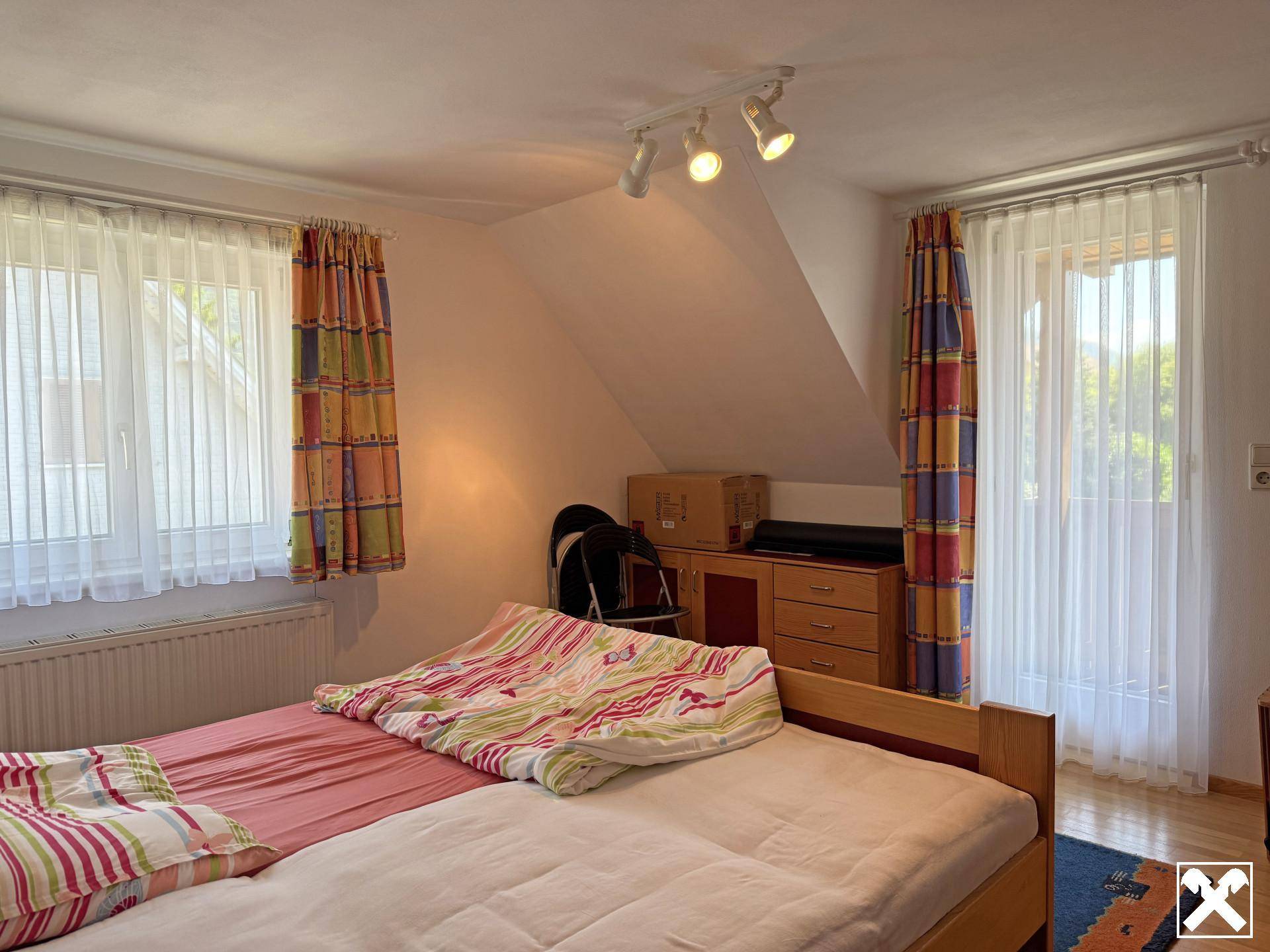 Schlafzimmer im Dachgeschoss mit schrägen Wänden, zwei Fenstern und farbenfrohen Vorhängen.