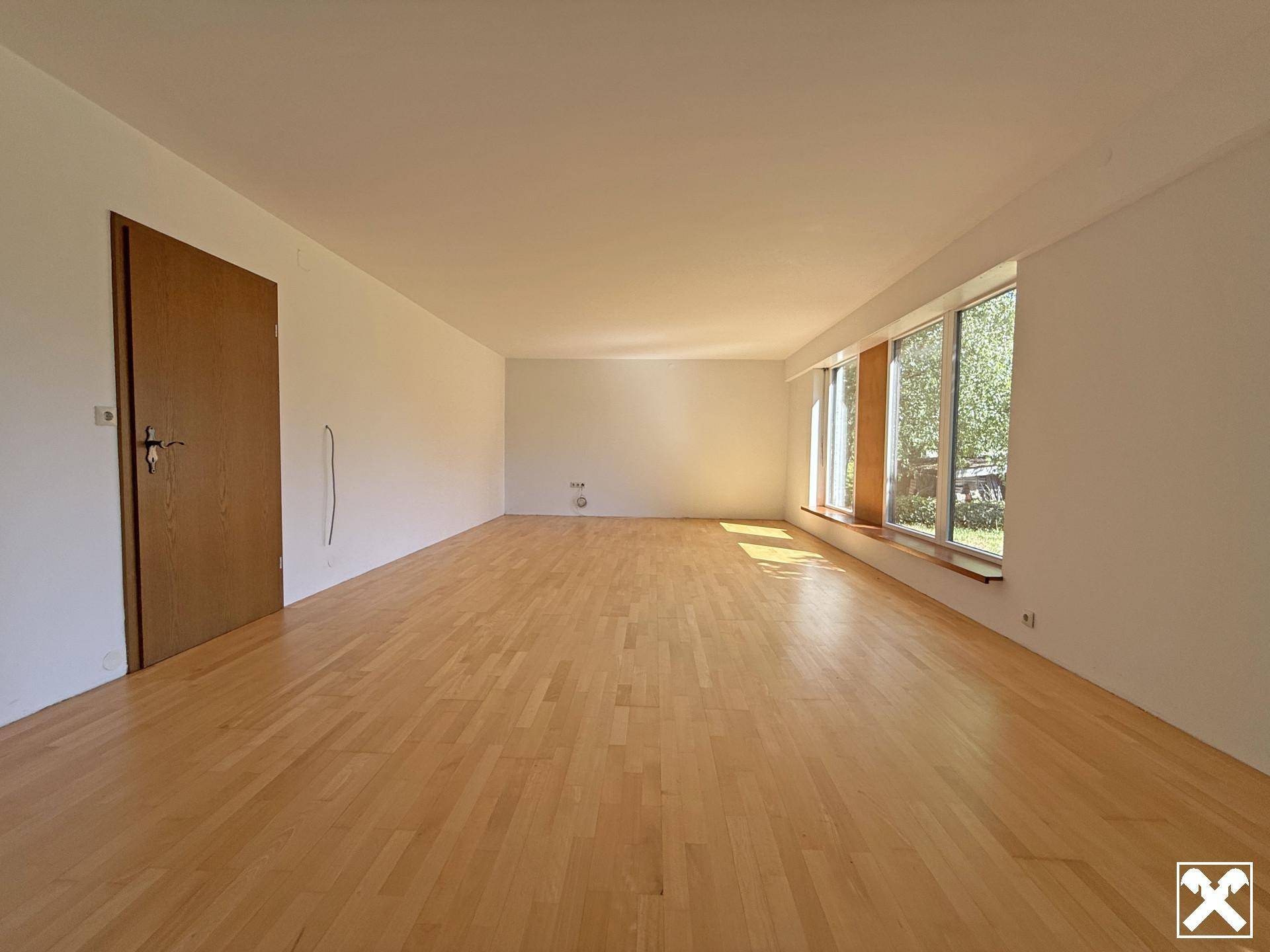 Geräumiges Wohnzimmer mit hellem Parkettboden und großen Fenstern, die viel Tageslicht hereinlassen.