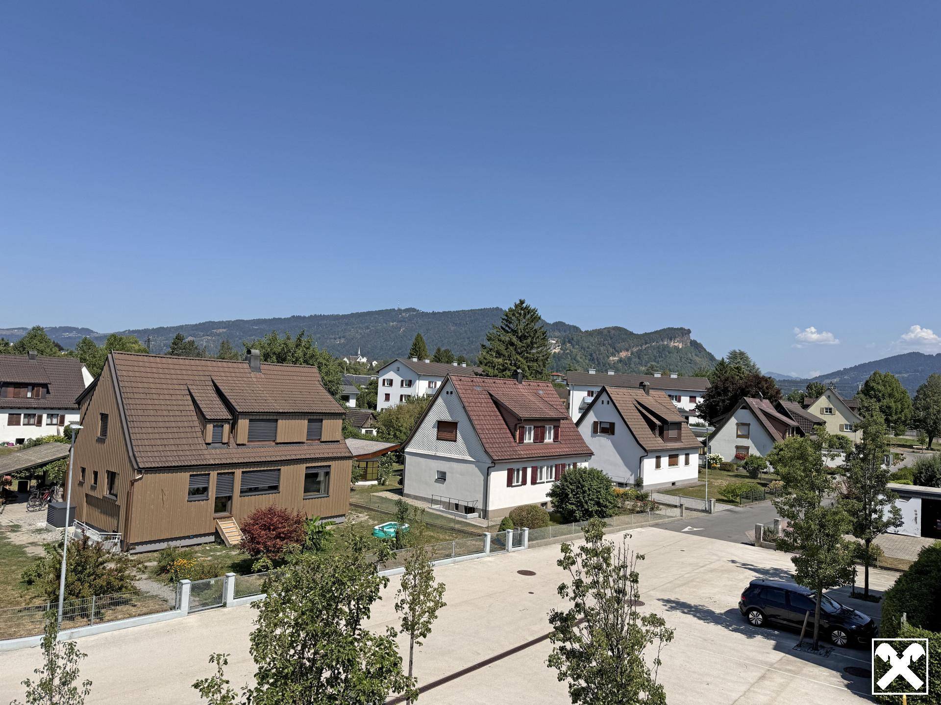 Blick über eine sonnige Wohnsiedlung mit Einfamilienhäusern und Gärten vor einer beeindruckenden Bergkulisse.