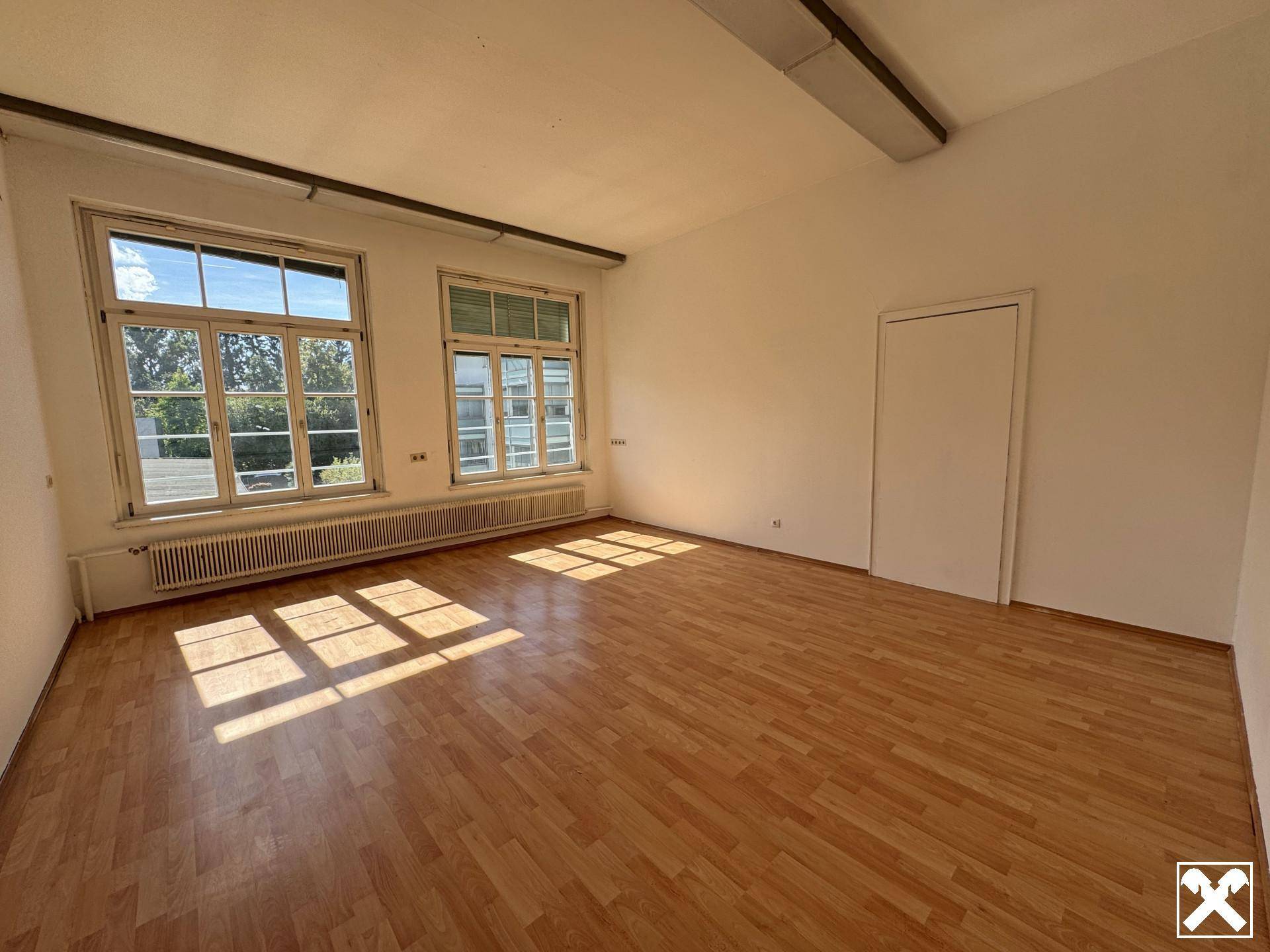 Heller Büroraum mit Holzparkettboden und großen Fenstern, die viel Tageslicht hereinlassen.