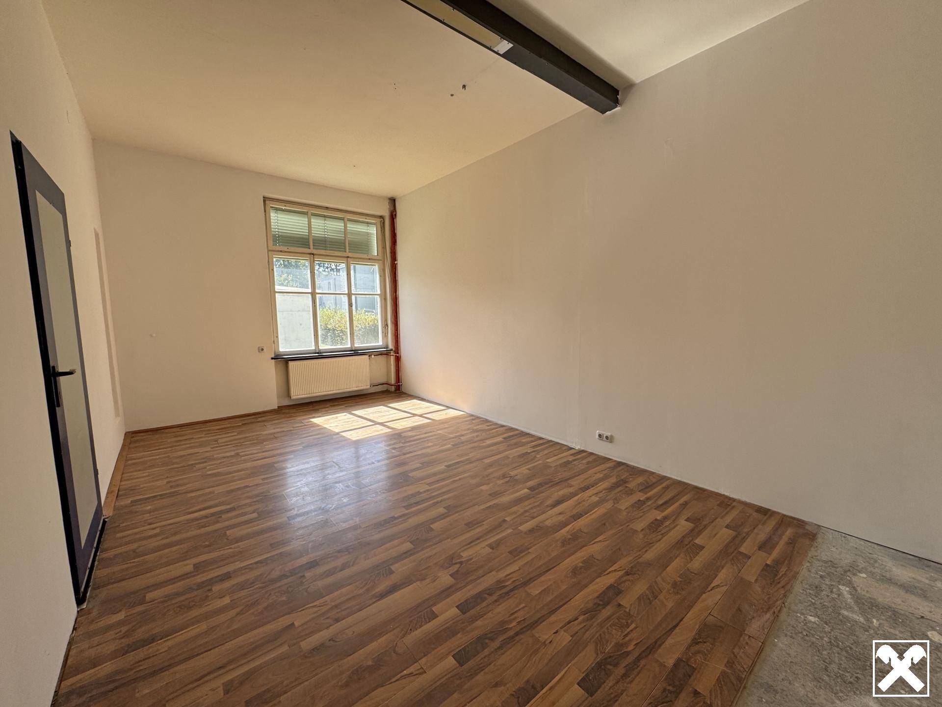 Leerer Büroraum mit Holzboden und einem Fenster, das den Raum mit natürlichem Licht versorgt.
