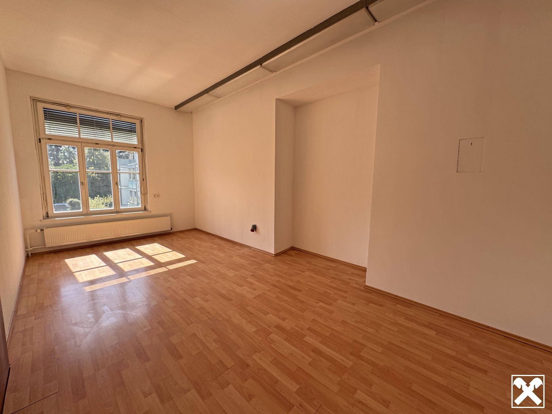 Heller Büroraum mit Holzparkettboden und einem großen Fenster, das viel Sonnenlicht hereinlässt.