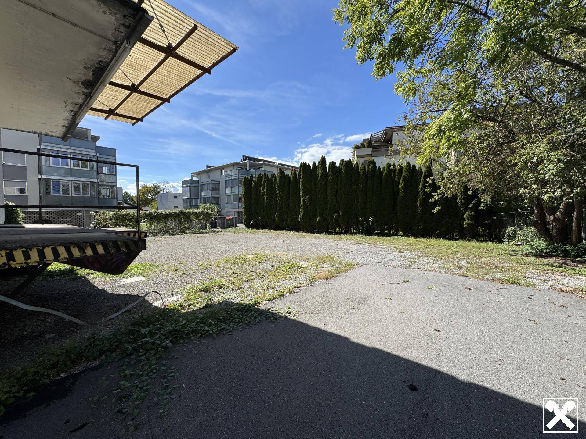 Freifläche neben einem Gebäude mit Hecke und Bäumen, umgeben von anderen Gebäuden.