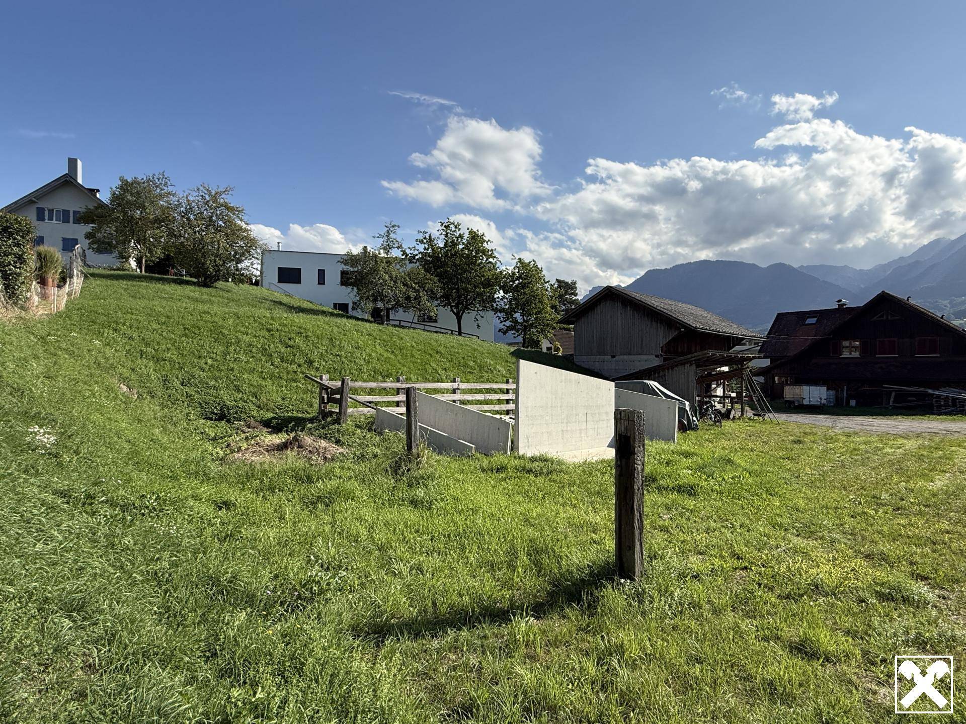 Hanglage mit grüner Wiese, umgeben von Wohnhäusern und einer Bergkulisse.