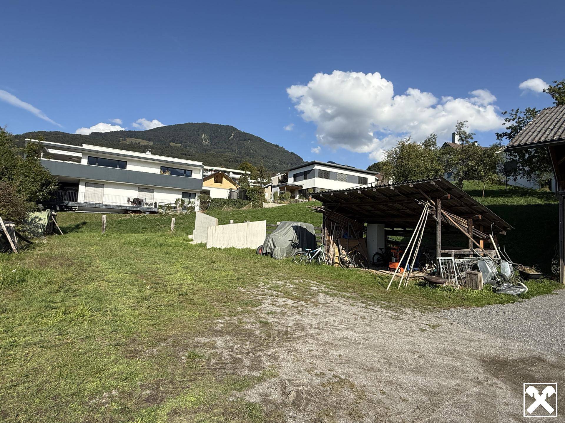 Ländliches Grundstück mit Holzschuppen und modernen Häusern im Hintergrund vor Bergpanorama.