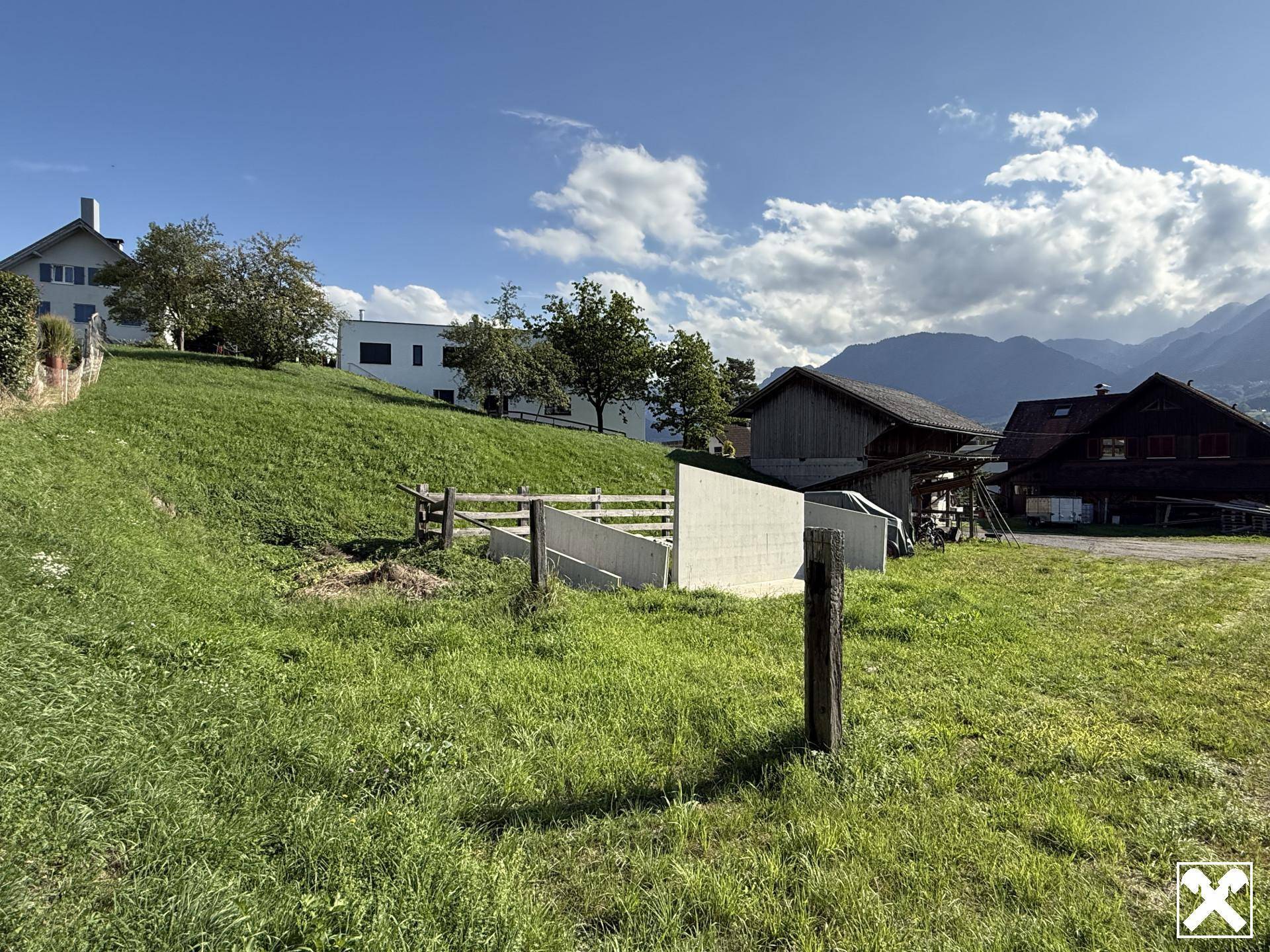 Grüne Wiese mit einer Betonstruktur, Holzzaun und traditionellen Gebäuden vor Bergkulisse.