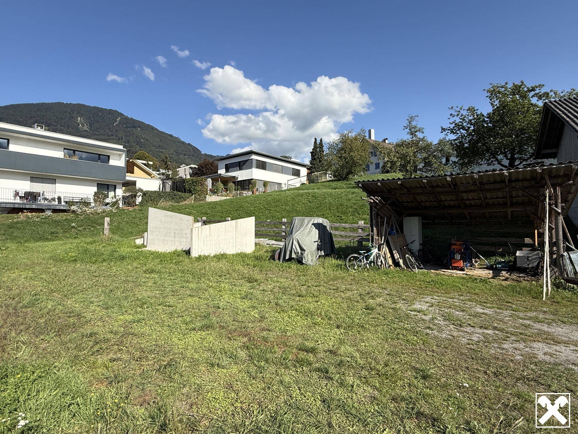 Grünfläche mit Betonwand, überdachtem Unterstand und modernen Häusern im Hintergrund.
