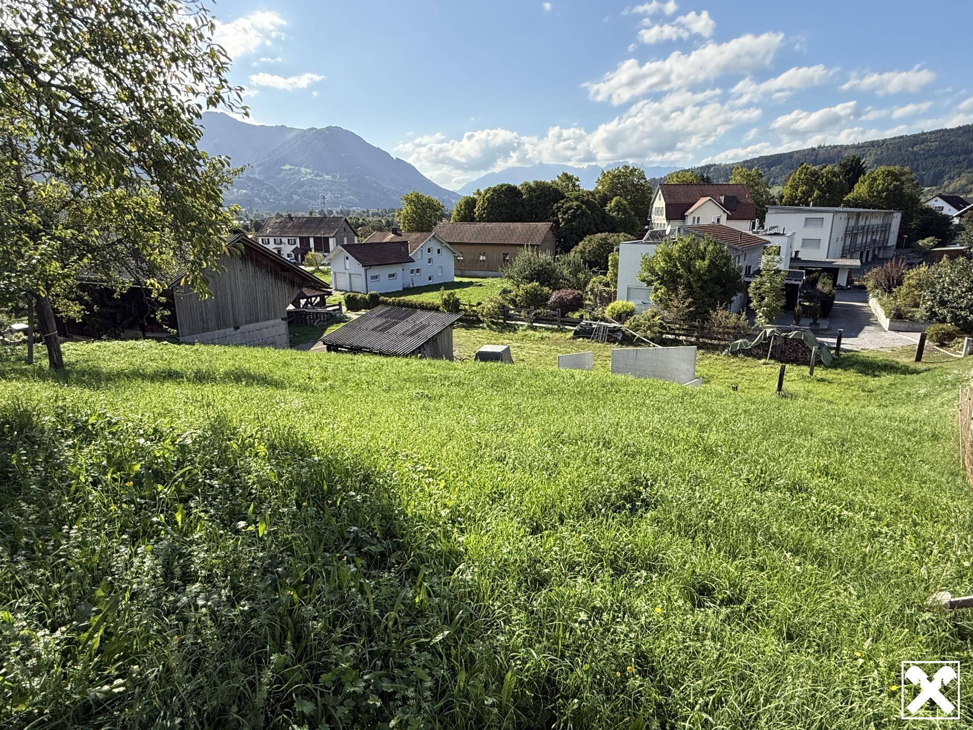 Panoramablick über eine grüne Wiese auf ein Dorf mit Häusern und die umliegende Berglandschaft.
