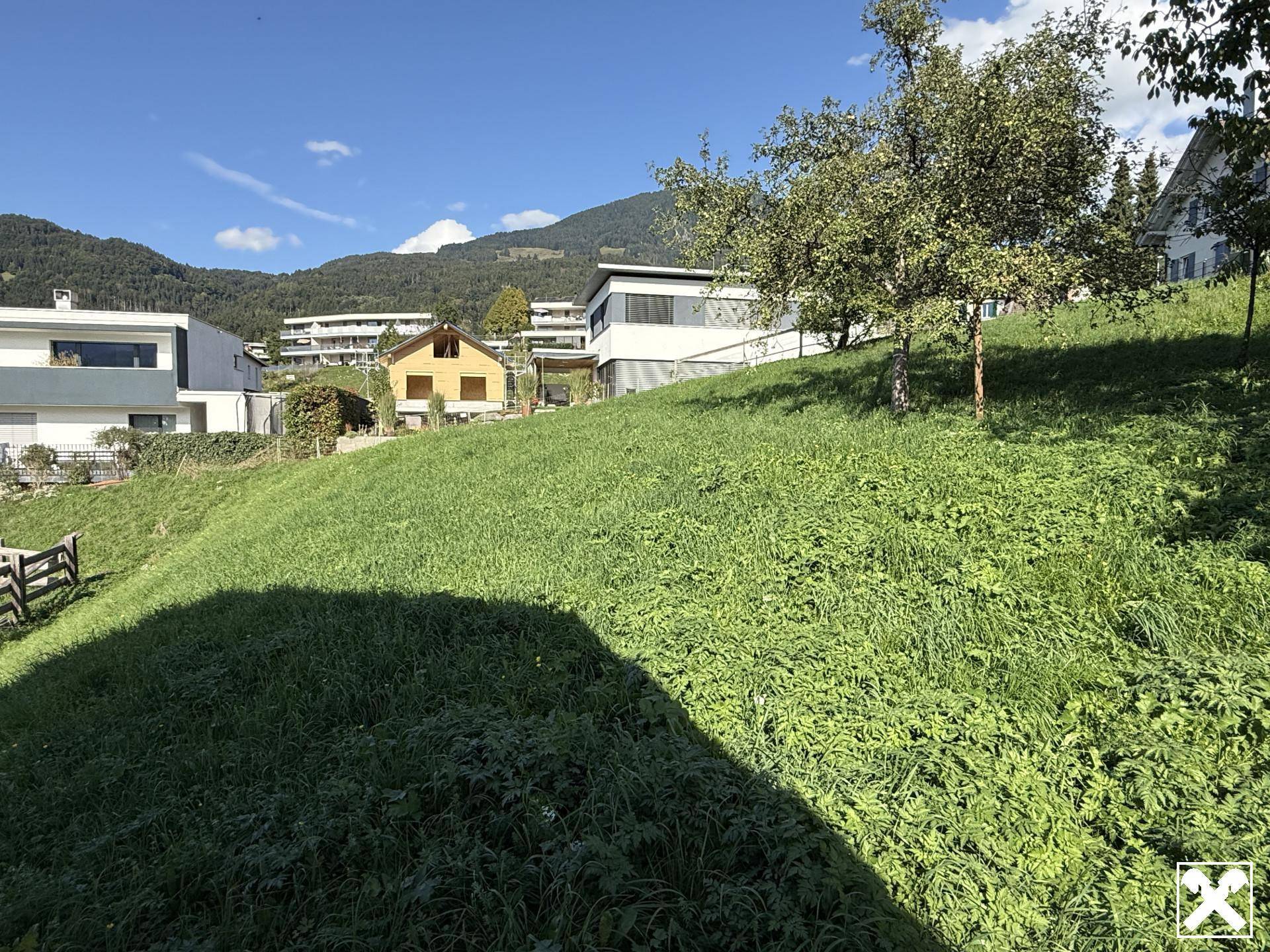 Weitläufige grüne Wiese mit verschiedenen Gebäuden und Bergen im Hintergrund an einem sonnigen Tag.