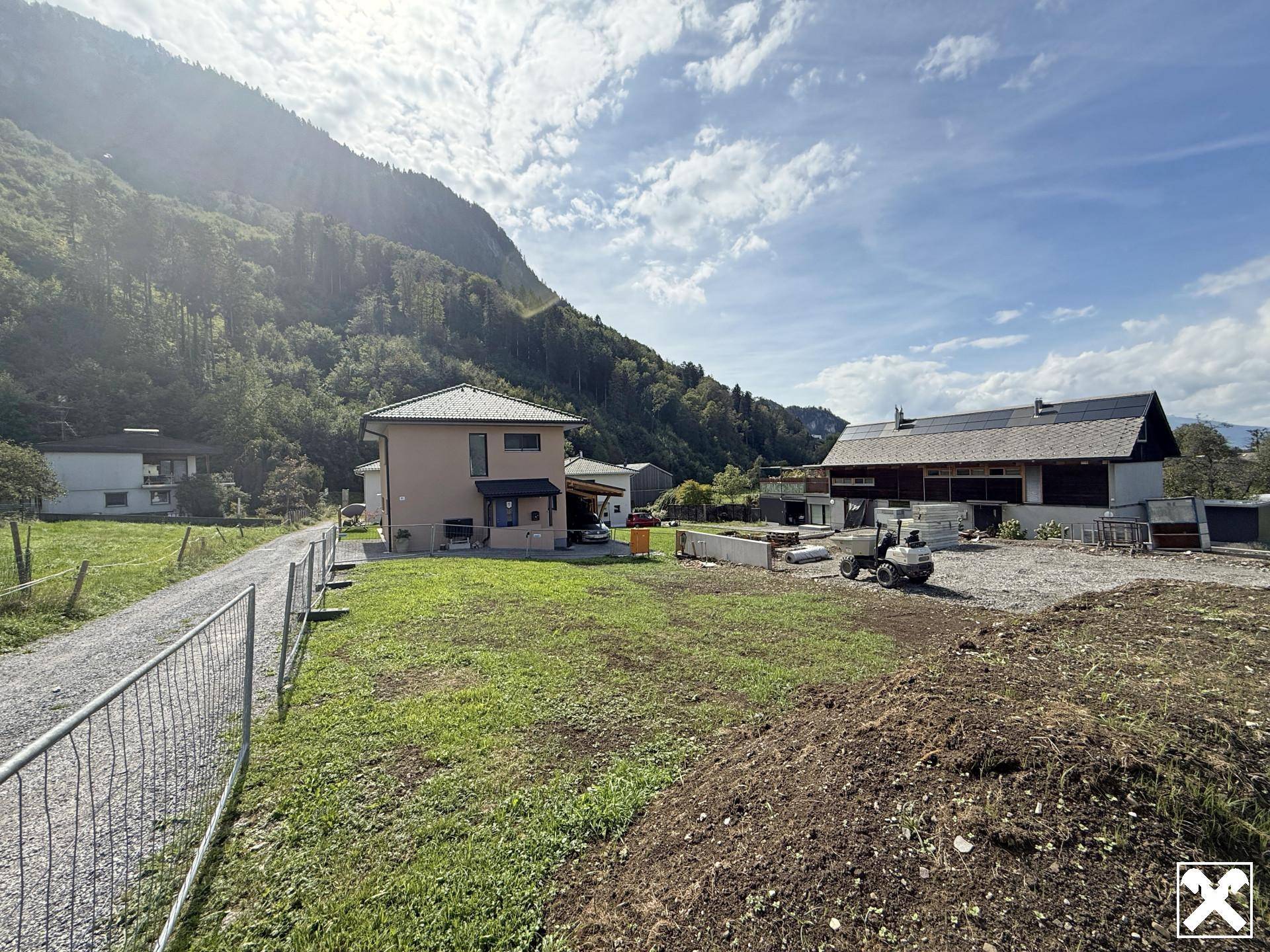 Weitläufiges Grundstück mit Neubau und Baustelle, umgeben von grünen Hügeln und Bergen unter blauem Himmel.