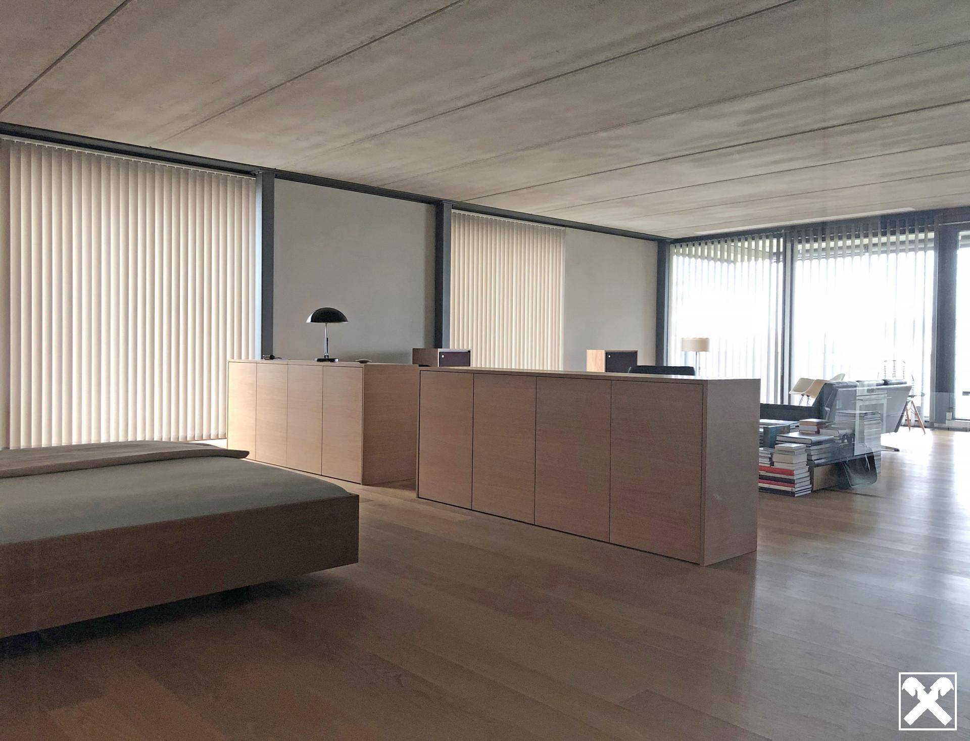 Helles Schlafzimmer mit Holzboden und großen Fenstern, die in einen offenen Wohnbereich übergehen.