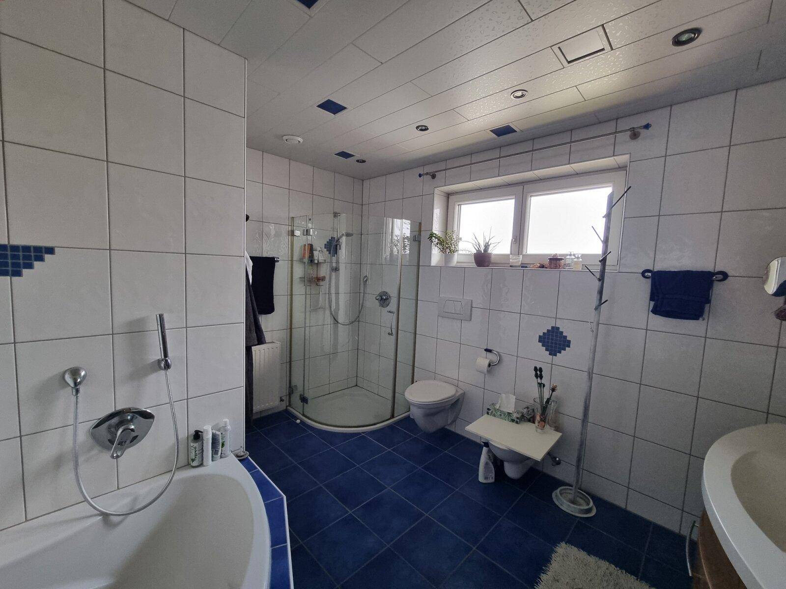 Badezimmer mit blauen Bodenfliesen, weißen Wandfliesen, Badewanne, Dusche und Fenster.