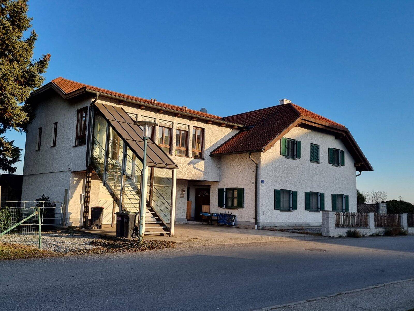 Vorderansicht des Hauses mit weißer Fassade, grünen Fensterläden und einem verglasten Treppenaufgang.