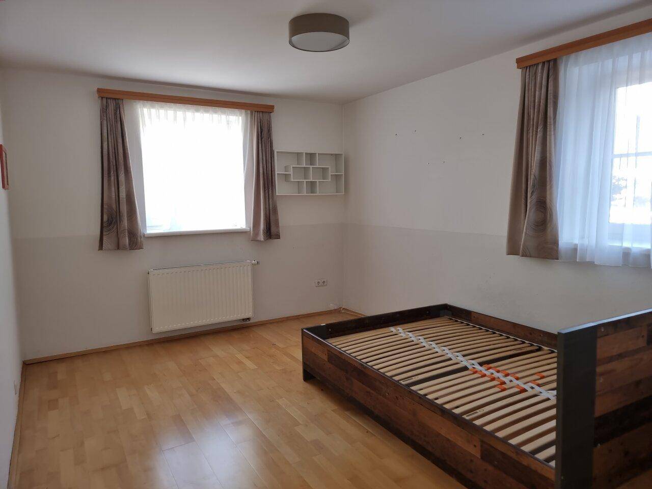 Helles Schlafzimmer mit Holzboden, großem Fenster und einfacher Einrichtung.