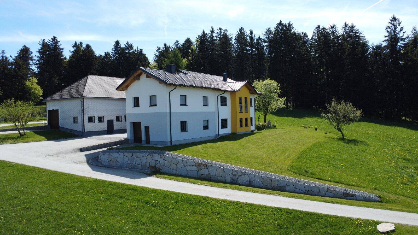 Modernes Wohnhaus mit angrenzendem Nebengebäude, umgeben von grünen Wiesen und Wald.