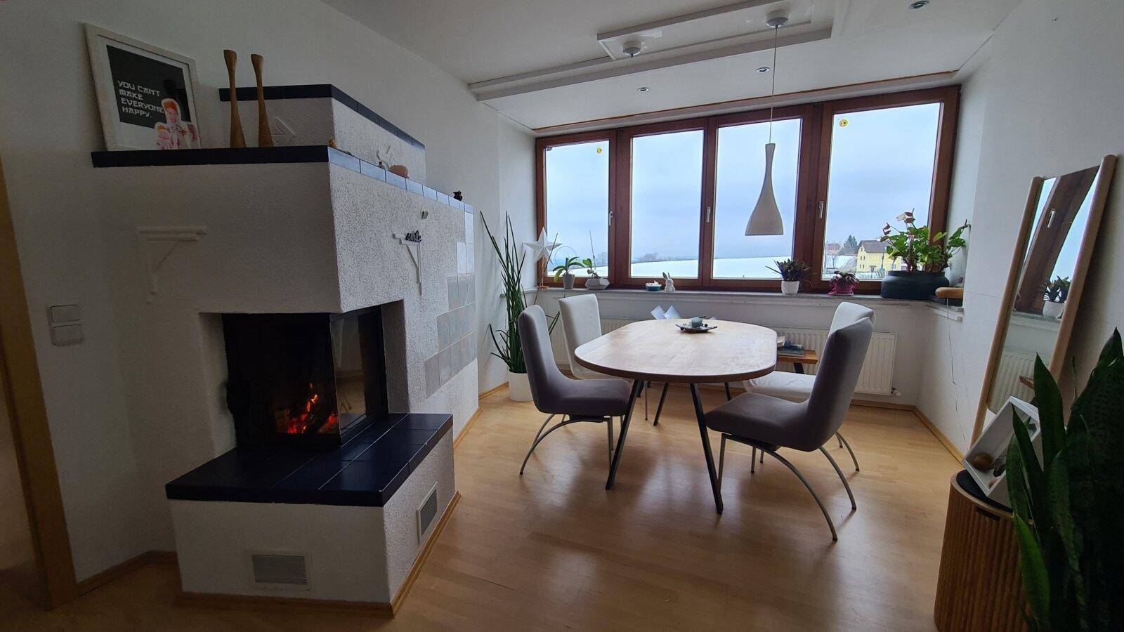 Gemütlicher Wohn- und Essbereich mit brennendem Kachelofen, Esstisch und großen Fenstern.